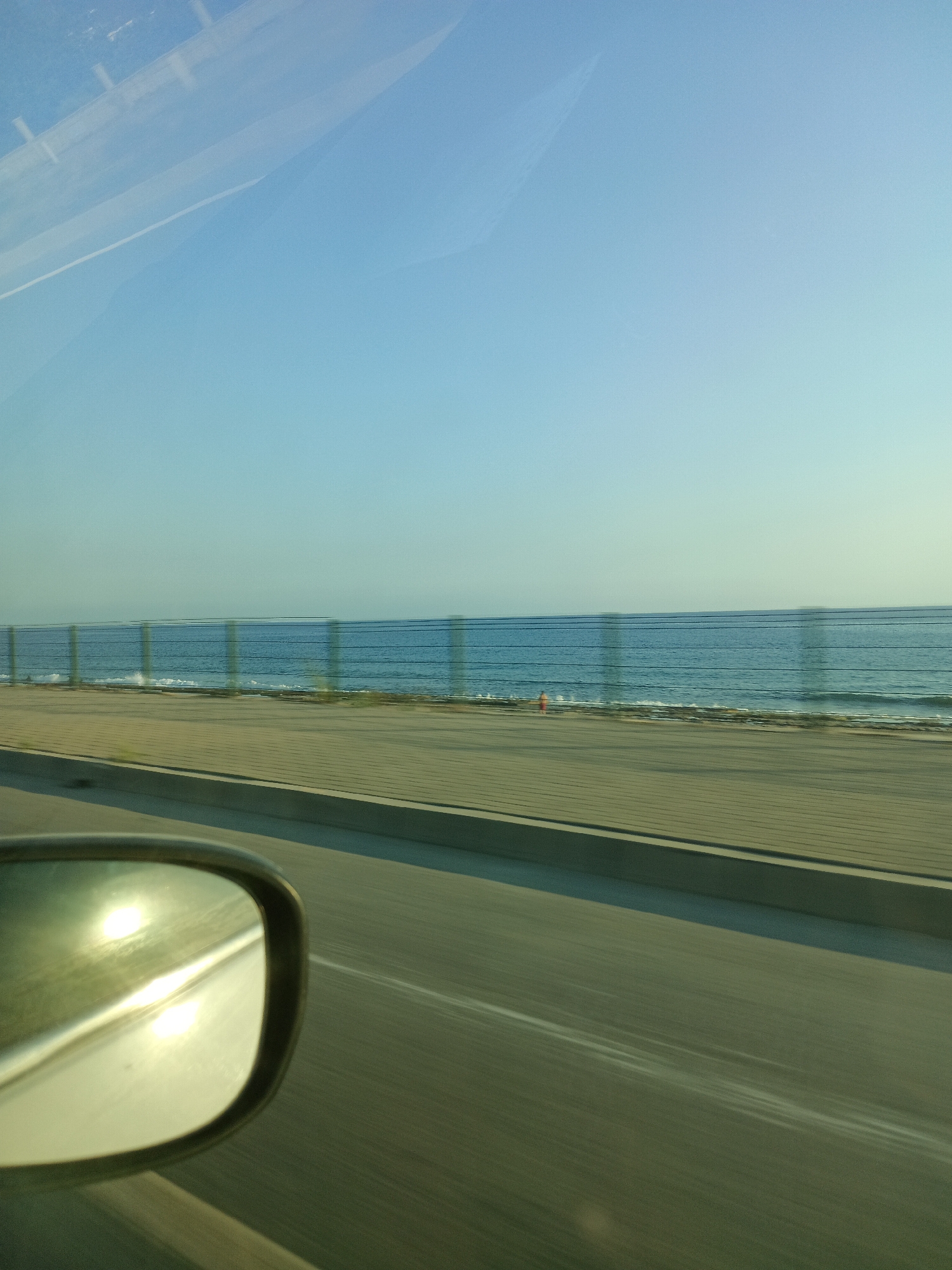 The image shows a view from a car window, likely on a coastal road in Alanya, Turkey. The scene is divided into three horizontal bands. The top band is a clear, pale blue sky. The middle band features the sea, a calm expanse of blue water with gentle waves breaking on a shore. A horizontal fence runs along the edge of the shore, obscuring some of the view. A small figure in an orange garment is visible on the beach. The bottom band shows a paved road and the side-view mirror of the car, reflecting the bright sunlight. The road appears to be a highway, indicated by the blurred lines and the sense of motion. The overall impression is one of a sunny day at the coast, viewed from a moving vehicle.