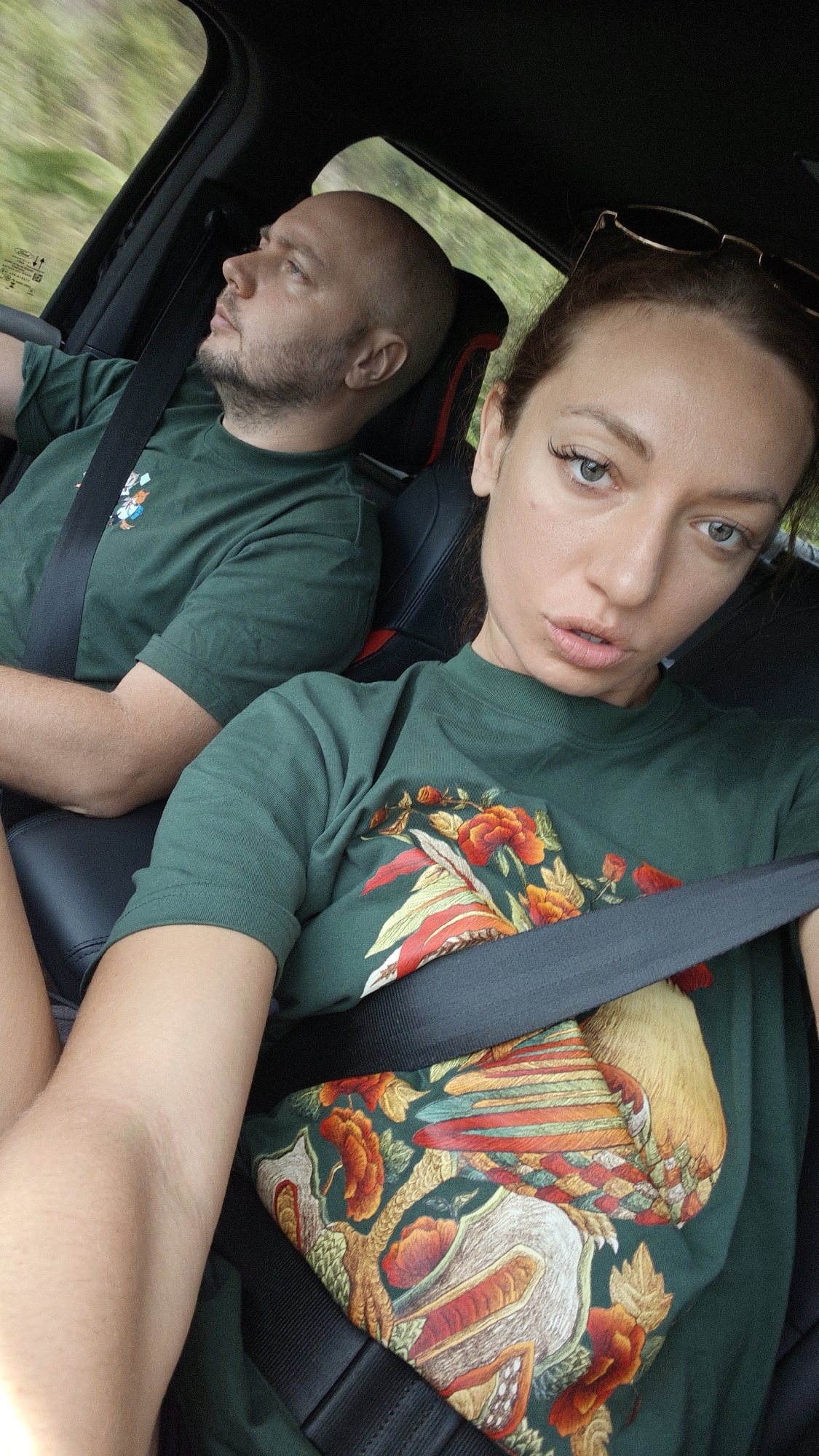 In a car interior, a selfie captures two individuals during what appears to be a daytime road journey, possibly in Duong Quy, Vietnam. A man, bald and with a short beard, occupies the driver's seat, looking thoughtfully towards the upper left. He wears a dark green t-shirt with a small graphic and has a black seatbelt fastened across his chest. Blurred green scenery is visible through the driver's window, suggesting movement. A small sticker on the window bears the "FORD" logo and other illegible text like "MAX LOAD."
In the foreground, a woman in the front passenger seat holds the camera, taking the picture. She has light eyes, long brown hair, and sunglasses perched on her head. Her expression is neutral to slightly pouty. She wears a dark green t-shirt adorned with a prominent, colorful design featuring a large bird, possibly a rooster or pheasant, surrounded by red and orange floral patterns. A black seatbelt is fastened across her. The car's interior features black seats, with a hint of red stitching visible on the driver's headrest. Both subjects are securely buckled, indicating they are in motion.