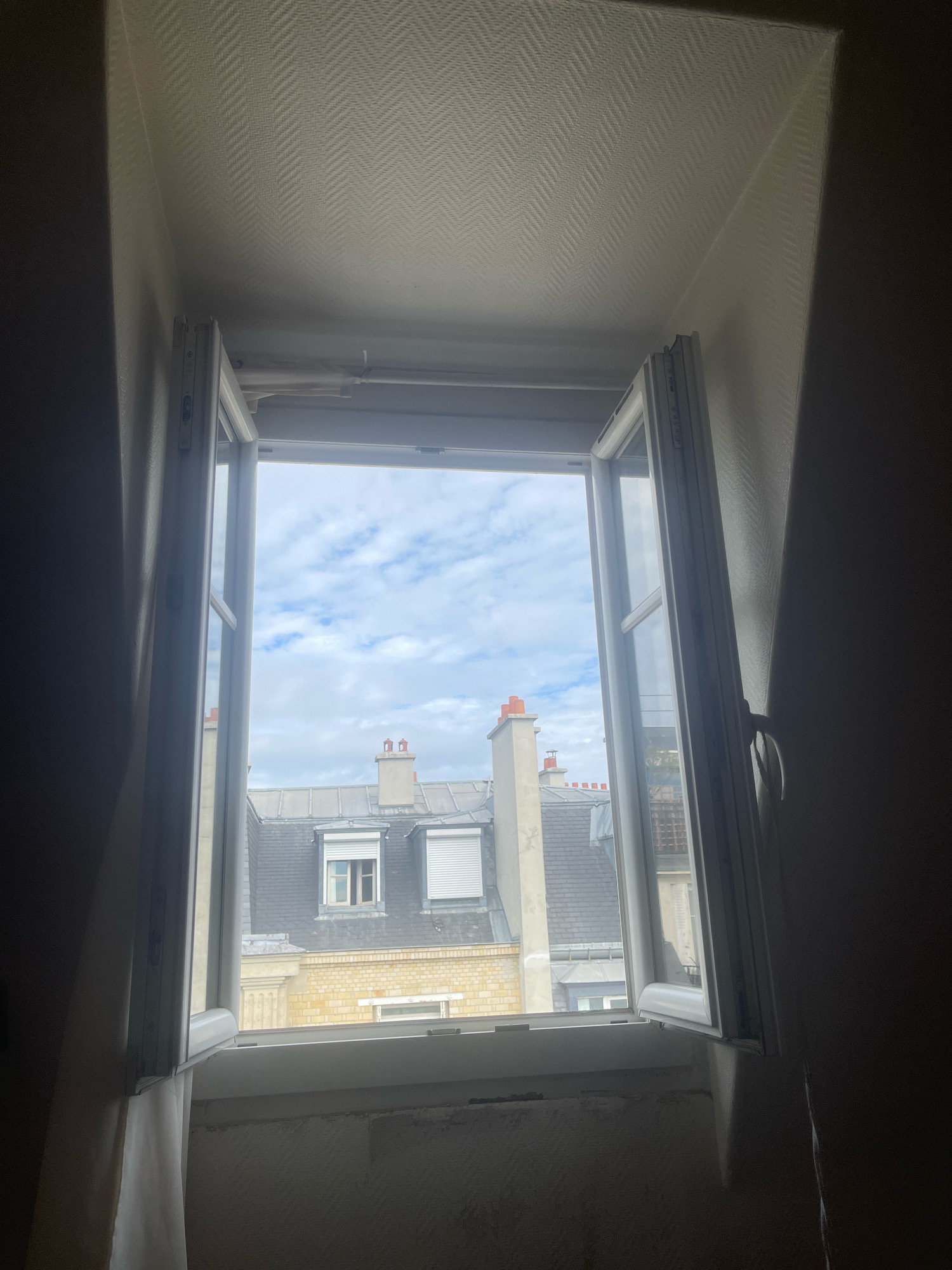 The image captures a view through an open window, revealing a Parisian cityscape. The window frame is white, and the window itself is slightly ajar, showing a sliver of the interior wall. To the left and right of the window, dark curtains hang down, obscuring the edges of the frame.
Outside the window, the sky is partly cloudy with patches of blue. Below the sky are the rooftops of buildings, characteristic of Paris. These rooftops are made of slate tiles, and several chimneys with terracotta caps are visible. Some buildings have dormer windows, and one has roller shutters. The architecture suggests a traditional Haussmannian style. The buildings appear to be made of stone and brick.
The immediate foreground, below the window sill, shows a rough, unfinished wall surface, indicating potential ongoing renovation or a lack of finishing. The ceiling above the window has a textured wallpaper, possibly a Herringbone pattern. The lighting suggests it is daytime. There are no people or animals visible in the image. The perspective is from inside a room, looking outwards.