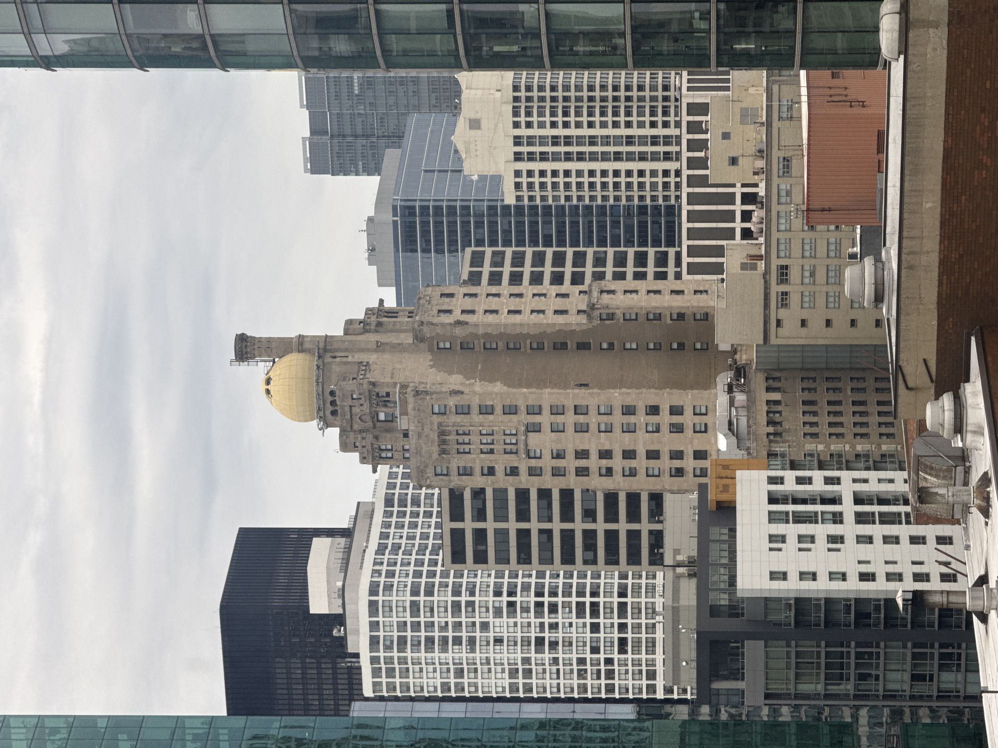 This is a vertical, eye-level shot of a dense urban cityscape in Chicago, USA. The scene is dominated by a variety of modern and older skyscrapers, creating a layered architectural landscape under an overcast sky. The image captures a mix of building materials, including glass, concrete, and brick, with sharp lines and geometric patterns defining the structures.
In the mid-ground, a striking building with a golden dome stands out, hinting at historical architecture amidst the contemporary designs. Surrounding it are numerous office buildings of varying heights and styles, from sleek glass facades to more traditional masonry structures with repetitive window patterns. One building features a unique, undulating white facade with a grid of windows. Another prominent structure is a dark, imposing building with a sharply angled facade.
The time of day appears to be overcast, with diffused light suggesting it is daytime, possibly morning or afternoon. The weather is cloudy, contributing to a muted color palette. There is no visible text in the image. The overall impression is of a bustling, metropolitan environment, characterized by its impressive and diverse architecture.