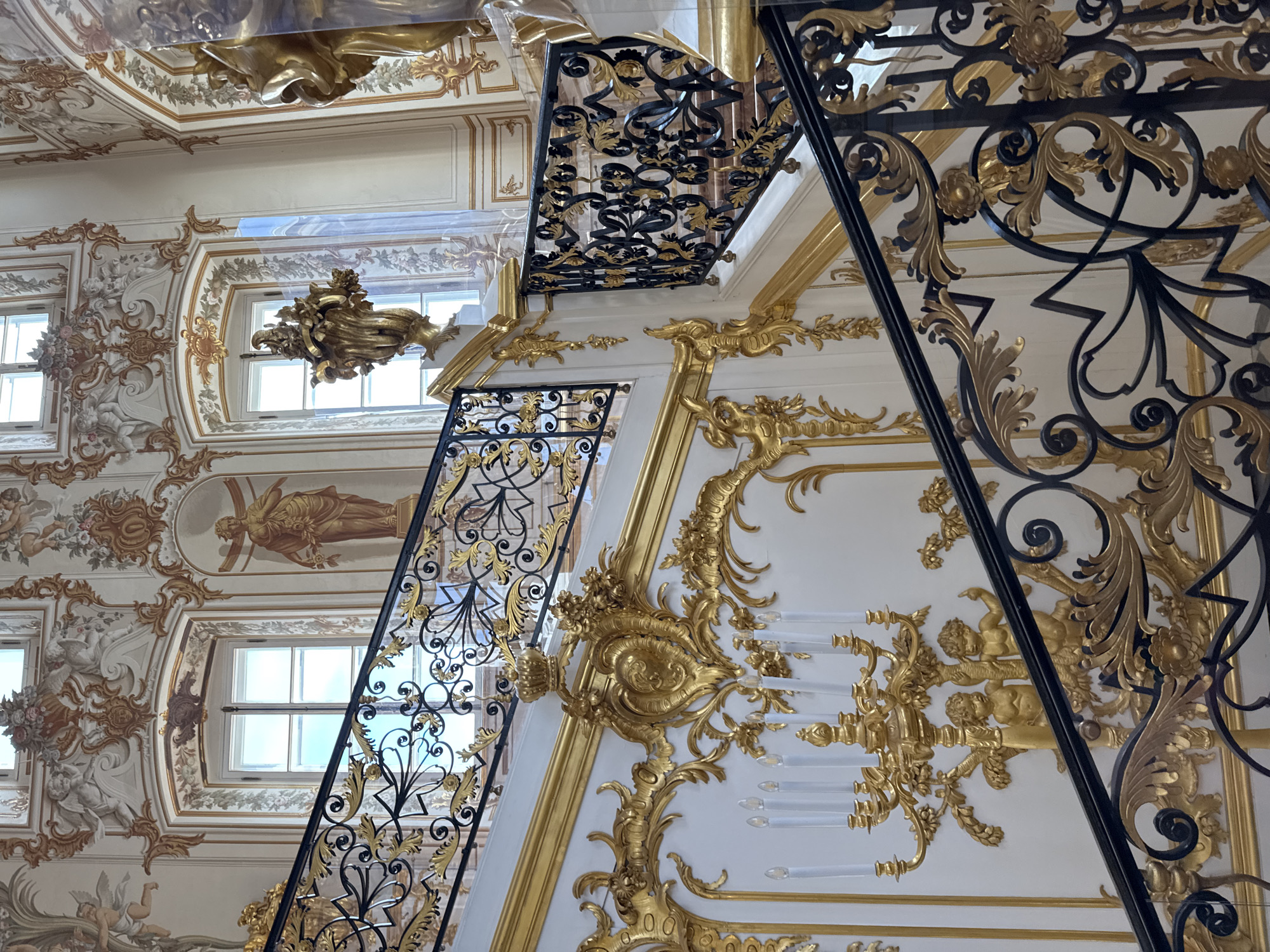The image shows a grand staircase in the State Rooms of the Grand Palace in Peterhof, Russia. The staircase is ornate, with black metal railings featuring gold leaf embellishments of scrolling foliage and floral motifs. The walls are decorated with intricate gold stucco work, including cherubic figures, floral swags, and cartouches, against a white background. Two large windows with multiple panes are visible in the background, allowing natural light to illuminate the space. The windows are framed by detailed gilded ornamentation and flanked by painted murals or stucco figures. A large, elaborate gold candelabra with multiple candle holders is mounted on the wall near the base of the stairs, adorned with gilded cherubs and foliage. The overall impression is one of opulent Baroque or Rococo architecture.