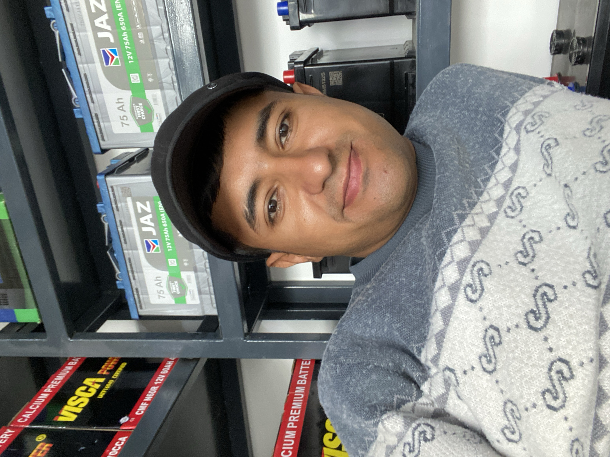 The image shows a man in Bukhara, Uzbekistan, posing in front of a shelf containing car batteries. The batteries are stacked on metal shelves. The brands visible are JAZ and VISCA. The man is wearing a black cap and a light-colored sweater with a repeating pattern. Some batteries indicate 12V 75Ah 650A (EN) and CMF N60R 12V 60AH specifications.