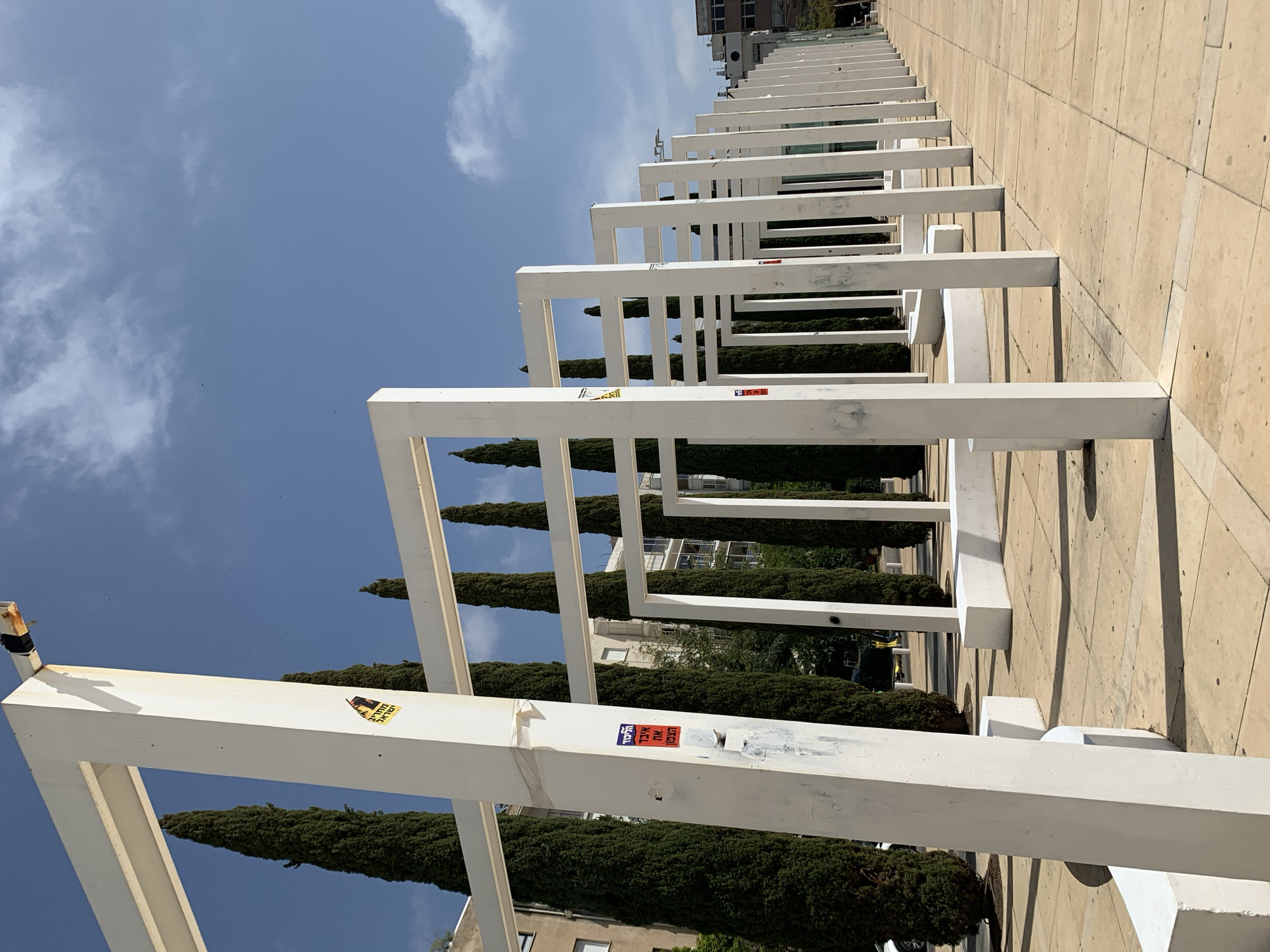 A horizontal-orientation, medium-distance shot shows the Dizengoff Square pergola in Tel Aviv, Israel, on a sunny day with a blue sky and some scattered clouds. The camera is positioned at street level and captures a perspective looking lengthwise down the pergola's structure.
The main subjects are the series of repeating white square frames that form the pergola. The frames are aligned in a row, receding into the distance. Between the frames, dark green, tall, conically shaped trees stand vertically, providing a contrast to the white frames and the blue sky visible through the openings. Stickers and some wear are visible on the frames closest to the camera.
To the right of the frames is a paved area consisting of light-colored, possibly limestone, blocks. The pavement shows signs of wear and is slightly uneven.
In the distance, indistinct buildings of varying heights are visible, providing a backdrop to the trees and pergola.
The environment is urban, and the lighting is bright and natural, casting shadows that emphasize the geometry of the structure.
