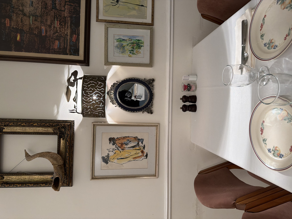 A wall displays various framed artworks, including a large painting, drawings, a decorative mirror, and a frame with a horn. Beside the wall, a formal table setting includes plates, silverware, glasses, and salt and pepper shakers. Two brown chairs are positioned on either side of the table.