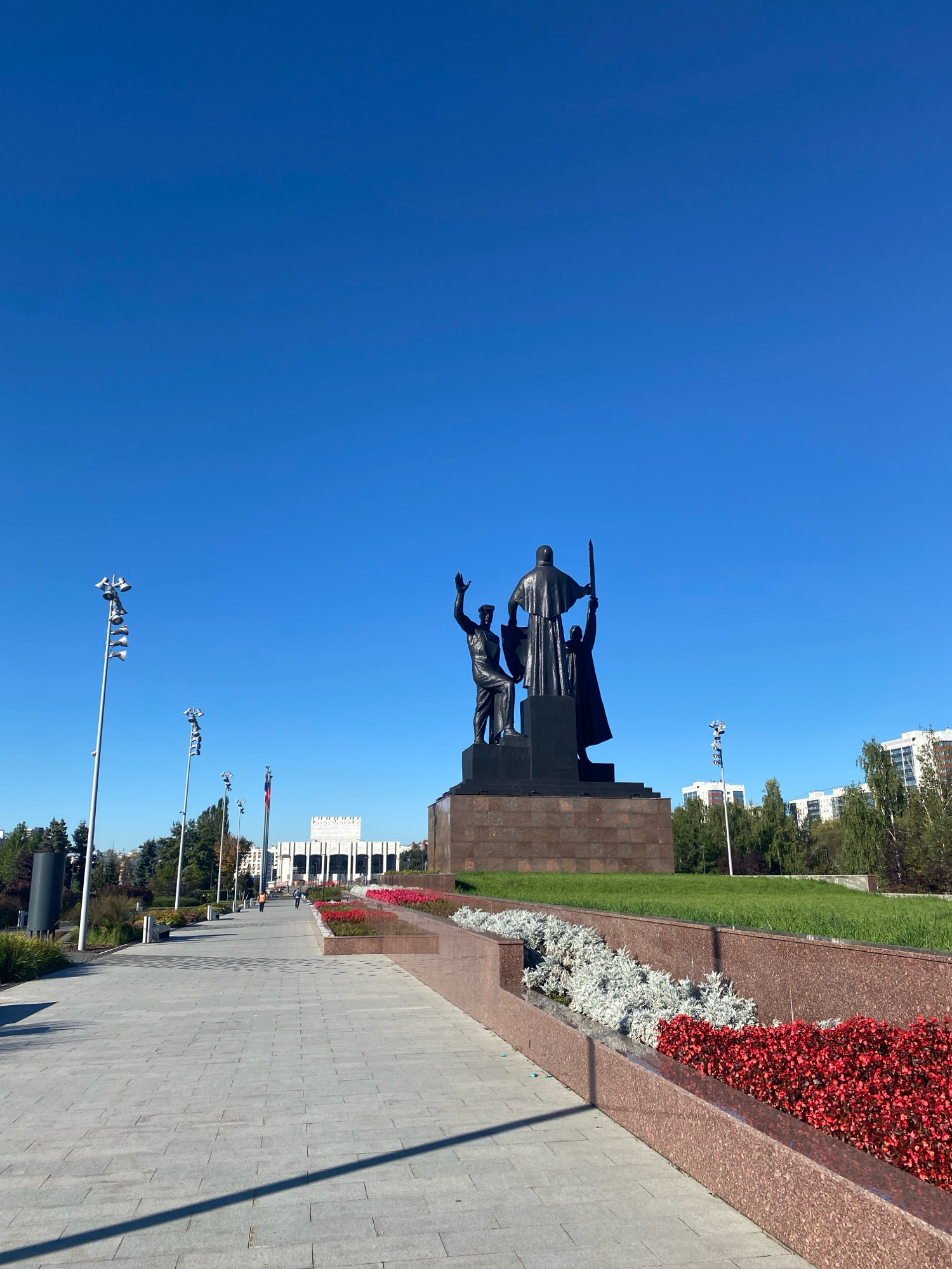The image shows a clear, blue sky above a wide, paved walkway. To the right, a large bronze monument stands on a stone pedestal, depicting three figures. The central figure, facing away from the viewer, holds a sword aloft, while two flanking figures are also in raised arm poses. In the background, a modern, white building with a colonnade is visible, flanked by trees and residential buildings. Several tall lampposts line the walkway on the left, and flower beds with red and white flowers are planted along the right side of the path. A shadow from the lamppost stretches across the walkway. The scene appears to be a public square or park in Perm, Russia, likely dedicated to a historical or national theme given the monumental sculpture and architecture.