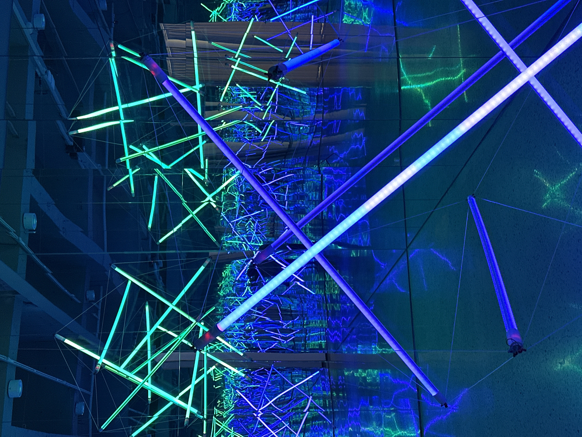 This image presents an abstract architectural lighting installation, likely indoors. The scene is dominated by an array of linear light fixtures, some glowing with a cool blue hue and others with a teal or greenish tinge. These fixtures are suspended by thin cables and arranged in a complex, intersecting pattern, creating a spatial geometric design. Reflections suggest the presence of mirrored surfaces, enhancing the illusion of depth and multiplying the light effects. The environment appears to be a contemporary or art-focused space. There are no visible people, actions, or interactions. It is not possible to confirm this image is located in Banan, China.