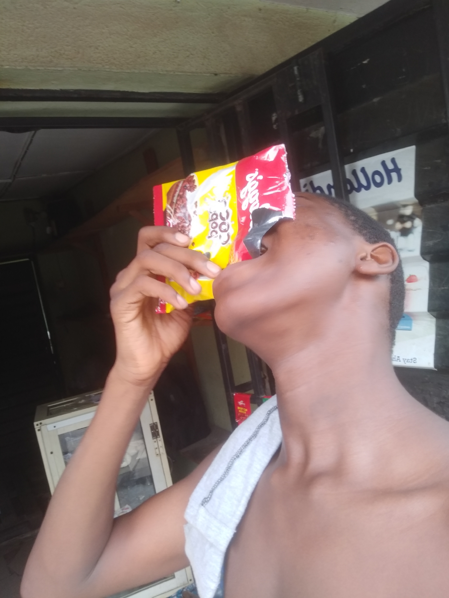 The image features a person, likely a young individual, consuming a snack or drink mix from a packaged item in what appears to be a shop or kiosk setting in Shangisha, Nigeria. The person is seen from the chest up in profile, with dark skin and short hair. Their head is tilted back, and a small, yellow and red packet is held to their mouth by their right hand, suggesting they are pouring its contents directly into their mouth.
The packet displays visible text, including "Coba Coba" (possibly a brand name related to cocoa) and "Jojo". The person is bare-chested or wearing a light-colored, possibly white, garment draped over their left shoulder.
The background is dimly lit, revealing structural elements of a building. To the right, a dark metal grate or window frame is visible. Behind the person's head, a sign with blue text is seen in reverse through glass, reading "HollwH" (possibly "Hollow" or part of a word) and "ya eA2" (likely "Stay A" or similar, reversed). To the left, a portion of a light-colored cabinet or display case with glass panels can be seen. The overall impression is an informal, semi-enclosed commercial space during daylight hours.