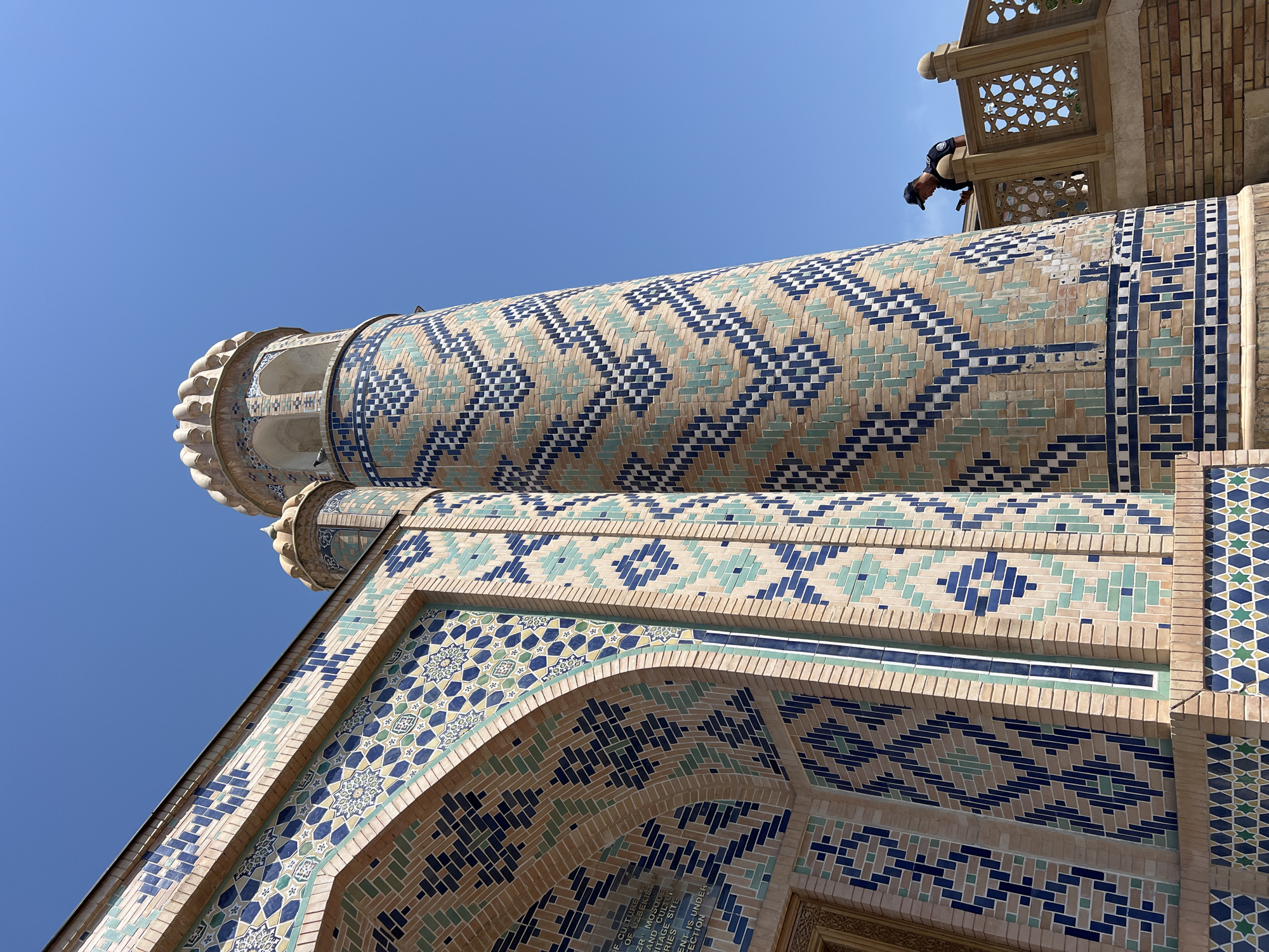 The image shows a low-angle view of a minaret and a portion of a mosque, both decorated with intricate geometric tilework. The minaret is cylindrical and features a decorative capital. The tile patterns are composed of small, rectangular tiles in shades of blue, green, and beige, creating diamond and zig-zag motifs. The mosque's facade also displays similar ornate tiling, with larger star-shaped patterns integrated into the design.
At the top of the image, a person wearing a cap and dark clothing is leaning over a balcony, appearing to look down. The person is silhouetted against a clear, bright blue sky, suggesting it is daytime and the weather is fair. The architectural style and vibrant tilework are characteristic of Islamic art and architecture found in Central Asia, with specific visual cues pointing to a location like Samarkand, Uzbekistan. Faint text is visible on a plaque near the bottom of the image, with legible words including "CULTURE," "UZBEKISTAN," "MOSQUE," and "HERITAGE SITE." The overall impression is one of grandeur and cultural significance, highlighting the detailed artistry of the building against a backdrop of a clear sky.