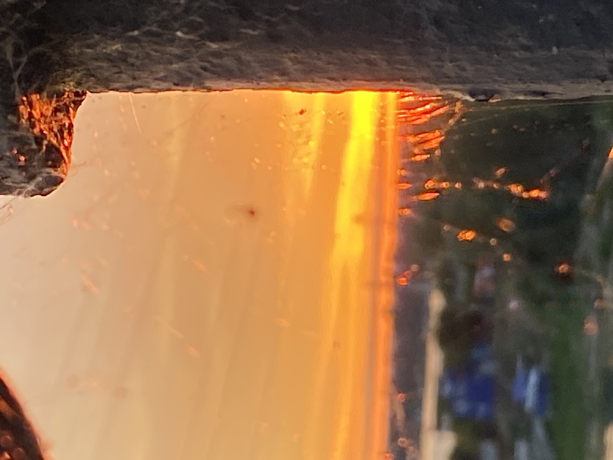 This is a photograph of a sunset viewed through a window, with a concrete pillar obscuring the right side of the frame. The sky is filled with vibrant orange and yellow hues, indicating the sun is low on the horizon. In the foreground, on the left side of the image, is a dark, blurred object, possibly part of the window frame, with some spiderwebs visible. Through the window, a distant, blurred landscape is visible, featuring some buildings and greenery, suggesting an urban or suburban setting. The overall impression is one of a dramatic sunset filtering through a somewhat obstructed view. The specific location in Novokuybyshevsk, Russia, is not discernible from the visual information provided in the image.