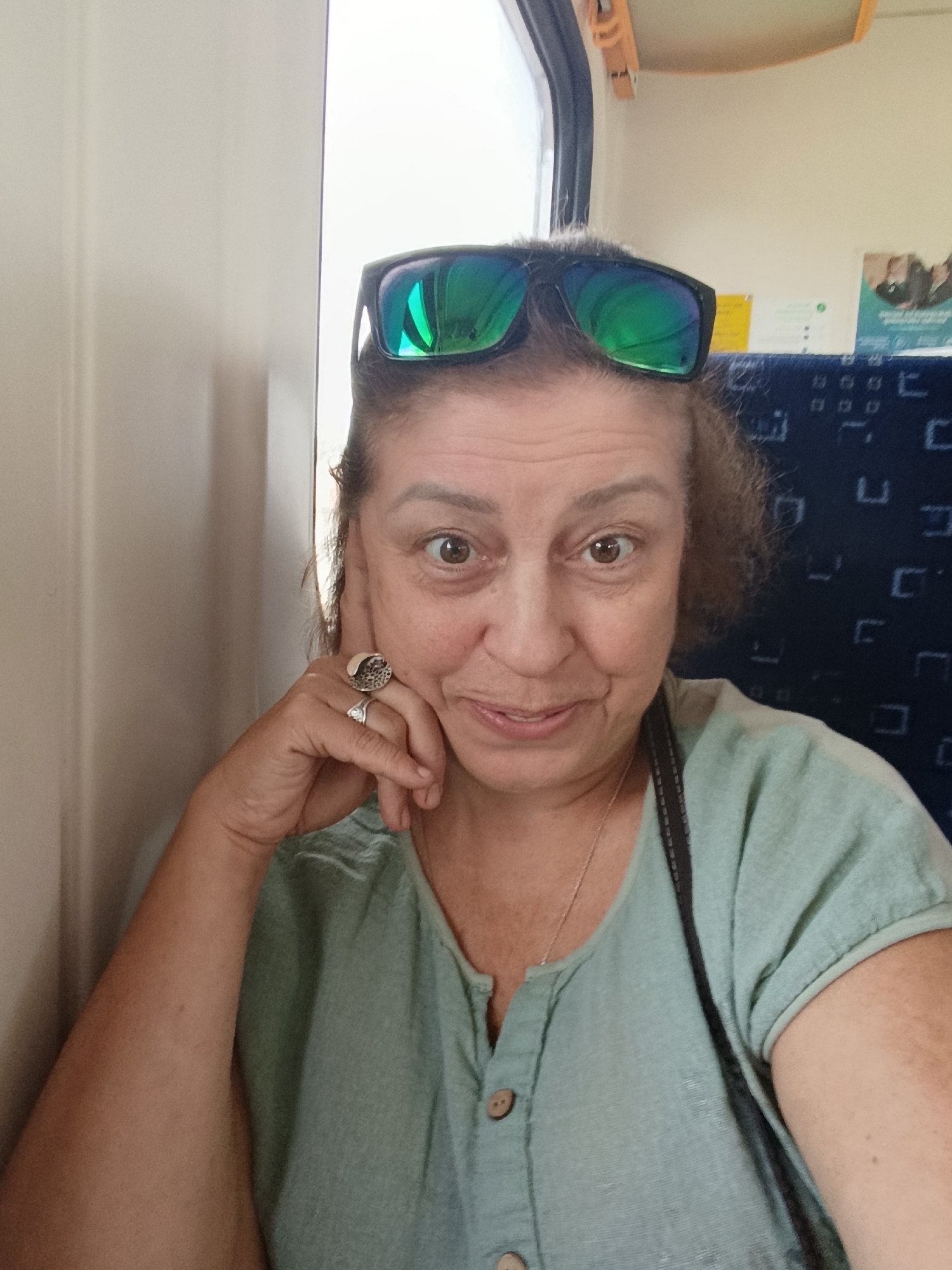 The image is a close-up, selfie-style photograph of a woman sitting on public transportation. The woman, who appears to be middle-aged, is looking directly at the camera with a slight smile. She is wearing a light green, button-up shirt and has sunglasses perched on top of her head, reflecting a green hue. She is wearing two rings on her left hand, which is resting on her cheek. The background shows a wall with a textured pattern, a window, and blue seats with a repeating geometric design, consistent with the interior of a train or bus. The environment appears to be indoors and during daylight. There is no visible indication of the location being in the city of Drezna, Russia.
