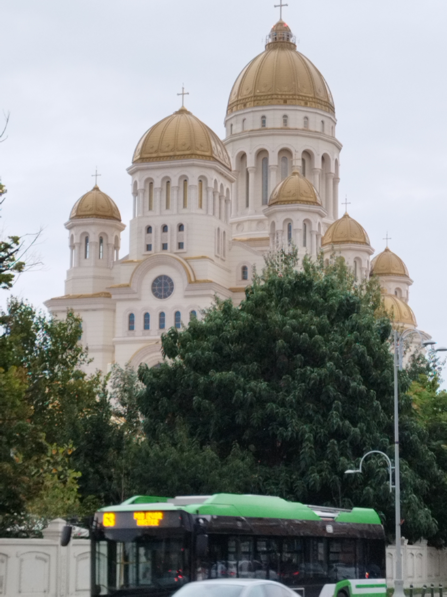 The image shows the Romanian Orthodox Cathedral in Bucharest, Romania, a prominent landmark characterized by its large, golden domes and white facade. In the foreground, a green and black bus is visible, its route display showing "63" and some unreadable text in yellow. The bus is in motion, suggesting a street scene in the city. Lush green trees partially obscure the view of the cathedral, indicating a setting with significant vegetation. The sky is overcast, contributing to a muted lighting condition. The overall scene depicts a typical urban environment with a juxtaposition of religious architecture and public transportation.