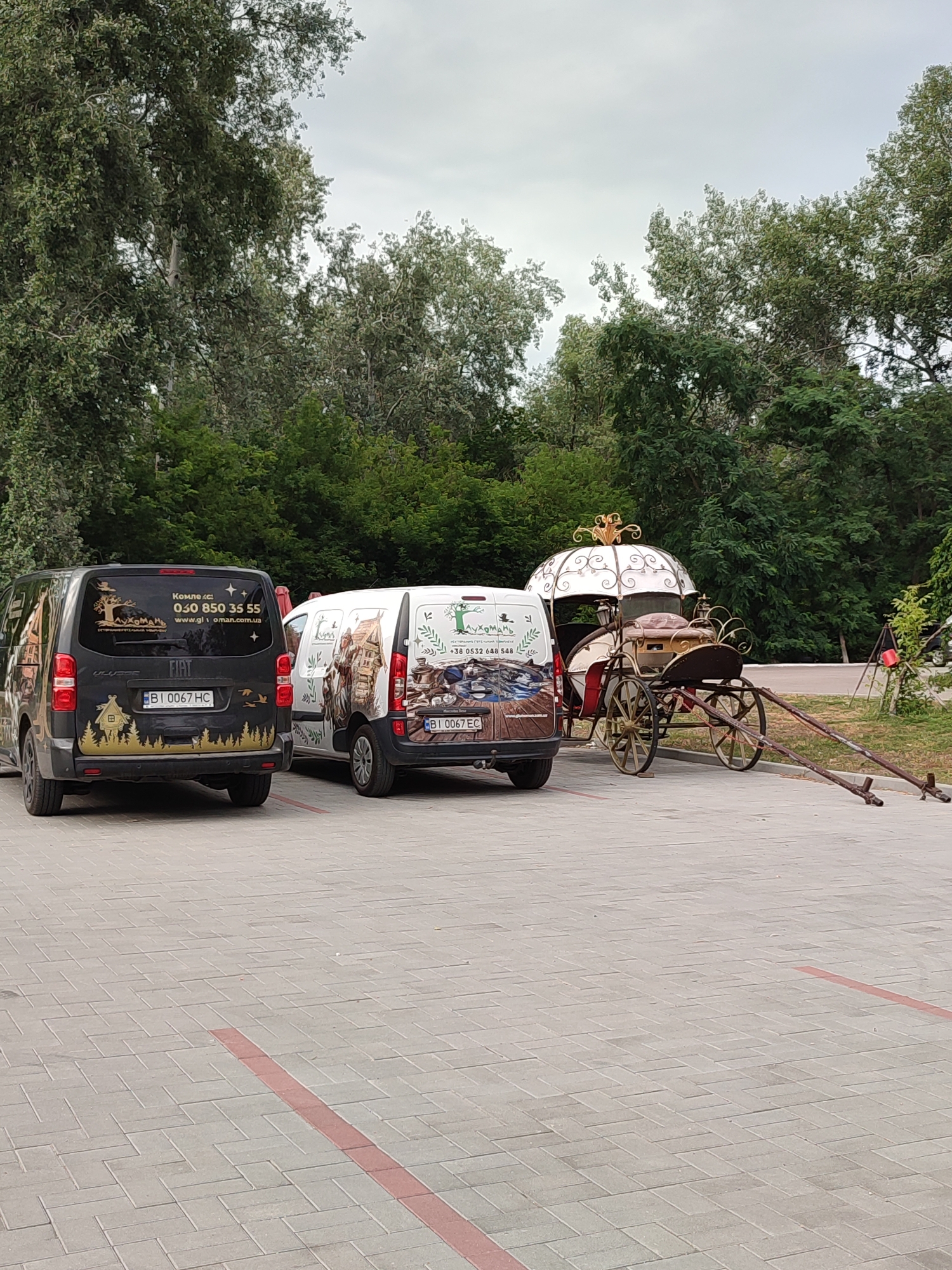 Two vans and a horse-drawn carriage are parked on a paved lot in Shcherbani, Ukraine. The van on the left is black and has a partial wrap with a silhouette of a forest and a house. The van on the right is white and has a full wrap depicting a pastoral scene with people in historical clothing. The horse-drawn carriage is ornate, with gold accents and a white canopy. It is positioned to the right of the white van. Trees line the background of the scene.