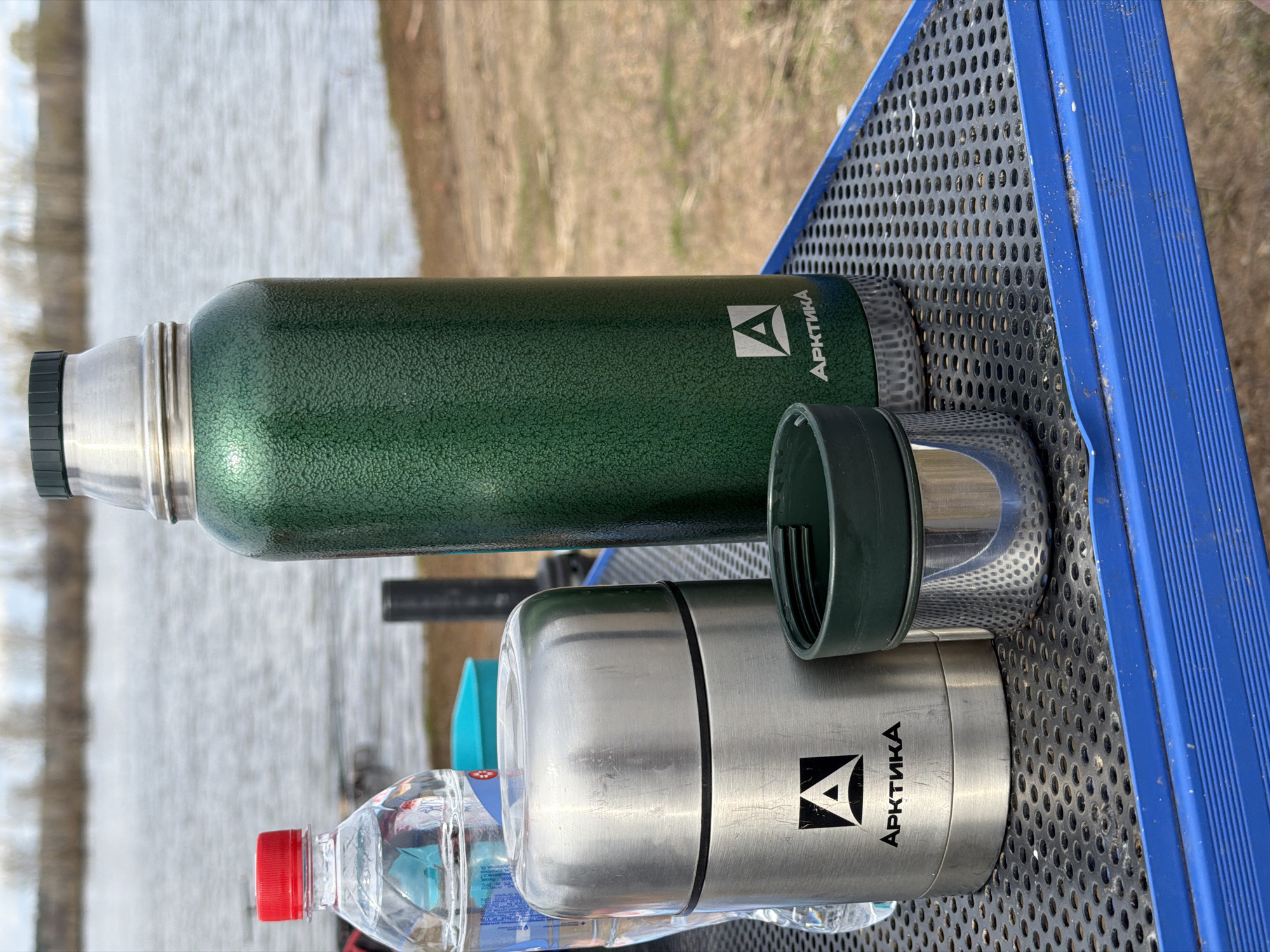 An outdoor shot captured in Nikolo-Beryozovka, Russia, features various beverages resting on a blue and black perforated metal surface, likely a picnic table or bench. In the foreground, a tall, dark green thermos with a textured finish stands upright. Its silver top and black screw cap are visible, while its matching cup is placed beside it. The green thermos bears the white Cyrillic text "АРКТИКА" (Arktika) and a stylized 'A' logo.
Next to it, a shorter, wider, brushed silver thermos also displays "АРКТИКА" (Arktika) in black Cyrillic letters with a black stylized 'A' logo, featuring a black band around its middle. Its silver cup is placed on top. A clear plastic water bottle with a red screw cap, filled with clear liquid, is positioned behind the silver thermos. The background shows a calm body of water, likely a river or lake, with faint ripples on its surface, and a blurred distant shore lined with vegetation under a bright, even daylight. The scene suggests a tranquil outdoor break or preparation for an activity by the water.