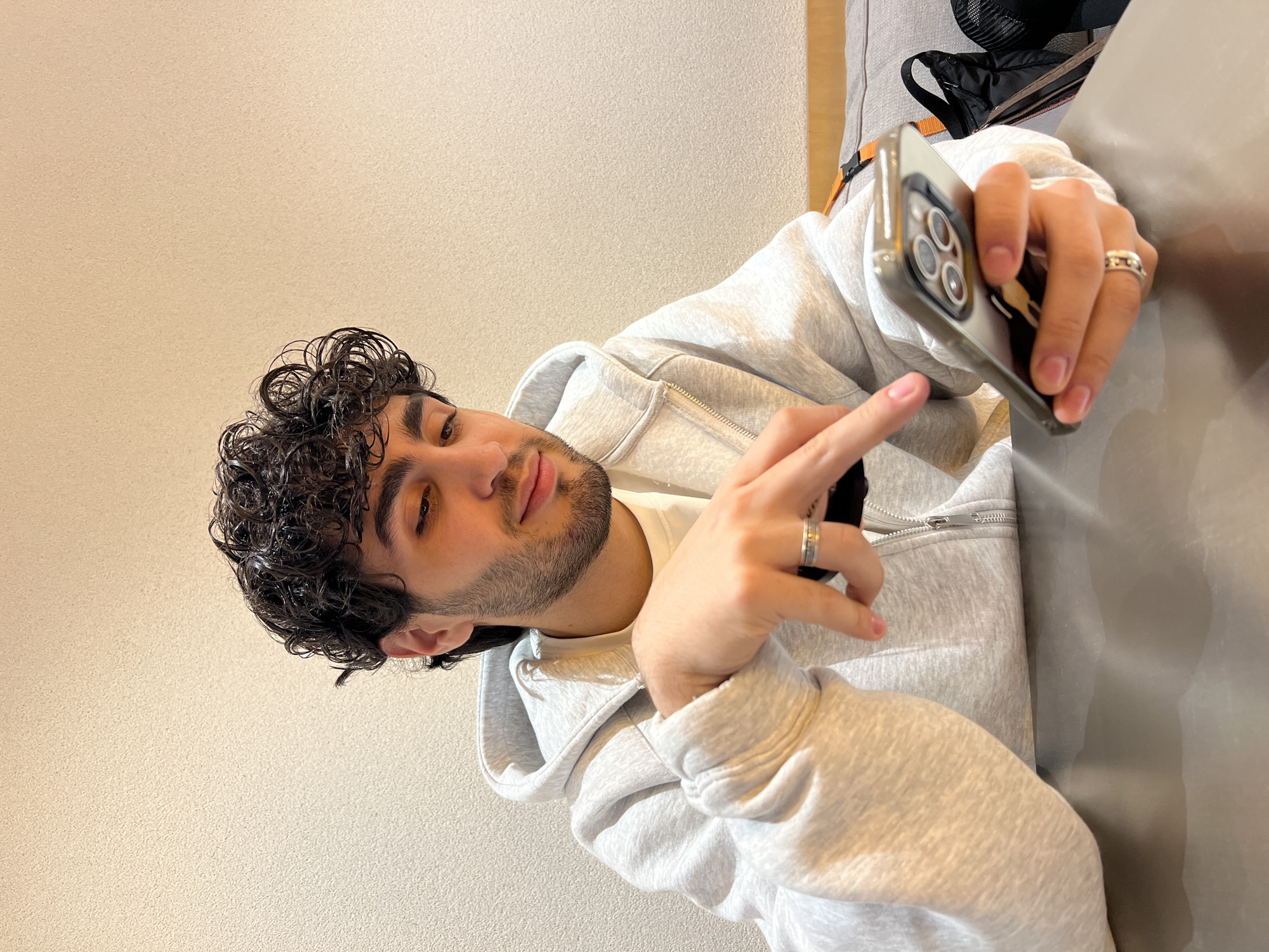 A medium shot depicts a young man with curly dark hair, a trimmed beard, and light skin wearing a light gray zip-up hoodie and a white t-shirt underneath. He is holding a cell phone in his right hand, and his left hand is raised with his index finger pointing towards the phone. The phone's rear camera array is visible. He is wearing rings on both ring fingers. A portion of a gray couch and a black bag are visible in the upper right corner. The background consists of a light-colored wall. There is no indication that this image was taken in Yerevan, Armenia.