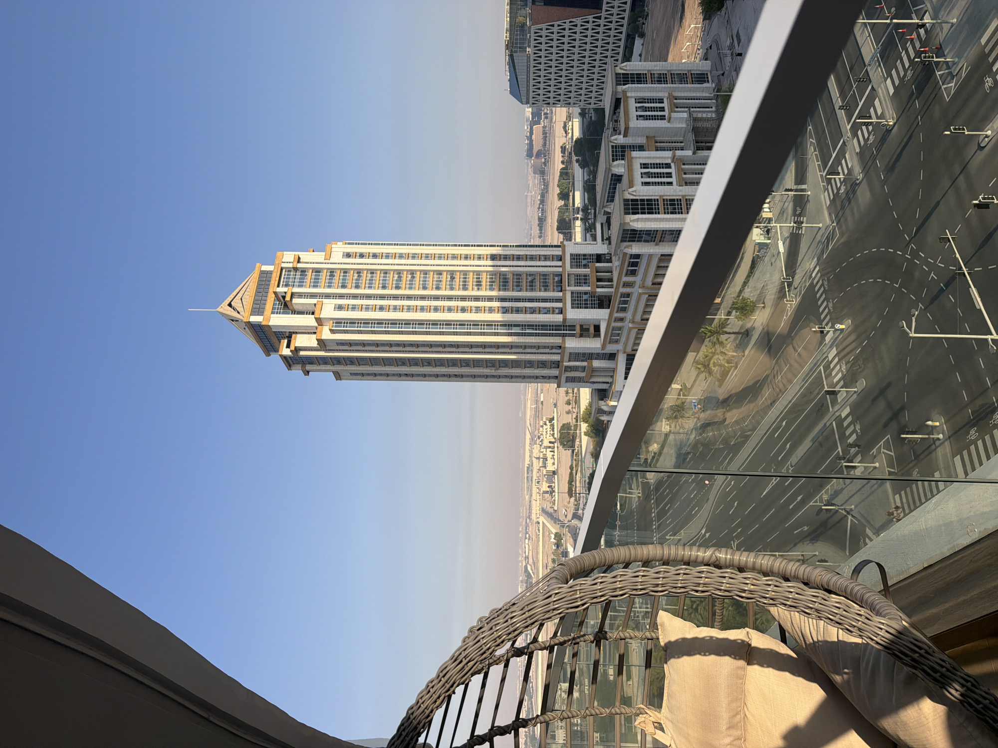 The image is an exterior, high-angle shot of a modern cityscape, taken from a balcony. In the foreground, a portion of a grey woven hanging chair with beige cushions is visible on the left, casting a shadow. The balcony railing, made of metal and glass, runs horizontally across the lower part of the frame. Beyond the railing, a multi-lane road with pedestrian crossings and traffic lights is visible, with cars and a few people on the pavement. Several buildings are present, including a tall, light-colored skyscraper with gold accents and numerous windows, positioned centrally in the mid-ground. To the right of this skyscraper is a lower, modern building with a distinctive geometric facade. In the background, the city stretches out with more buildings and a hazy horizon under a clear blue sky. The overall environment suggests a sunny day in an urban setting.