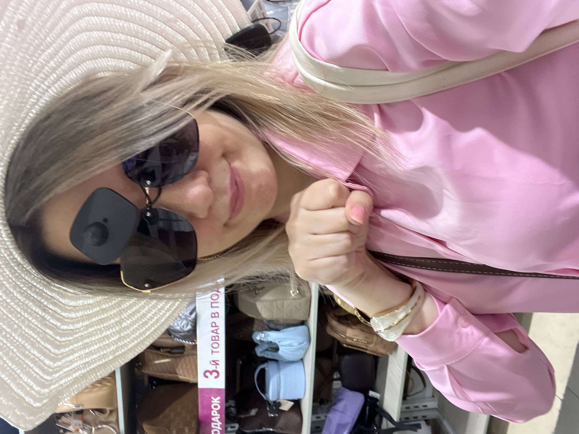 A blonde woman wearing a wide-brimmed straw hat and dark sunglasses poses for a selfie in what appears to be a store, possibly a department store in Sochi, Russia, given the text on a shelf sign in Cyrillic. The woman is wearing a bright pink collared shirt. Her right hand is clenched into a fist and held up near her face. A white shoulder bag strap is visible over her right shoulder. Behind her, shelves are stocked with various handbags and accessories. The lighting suggests an indoor retail environment.