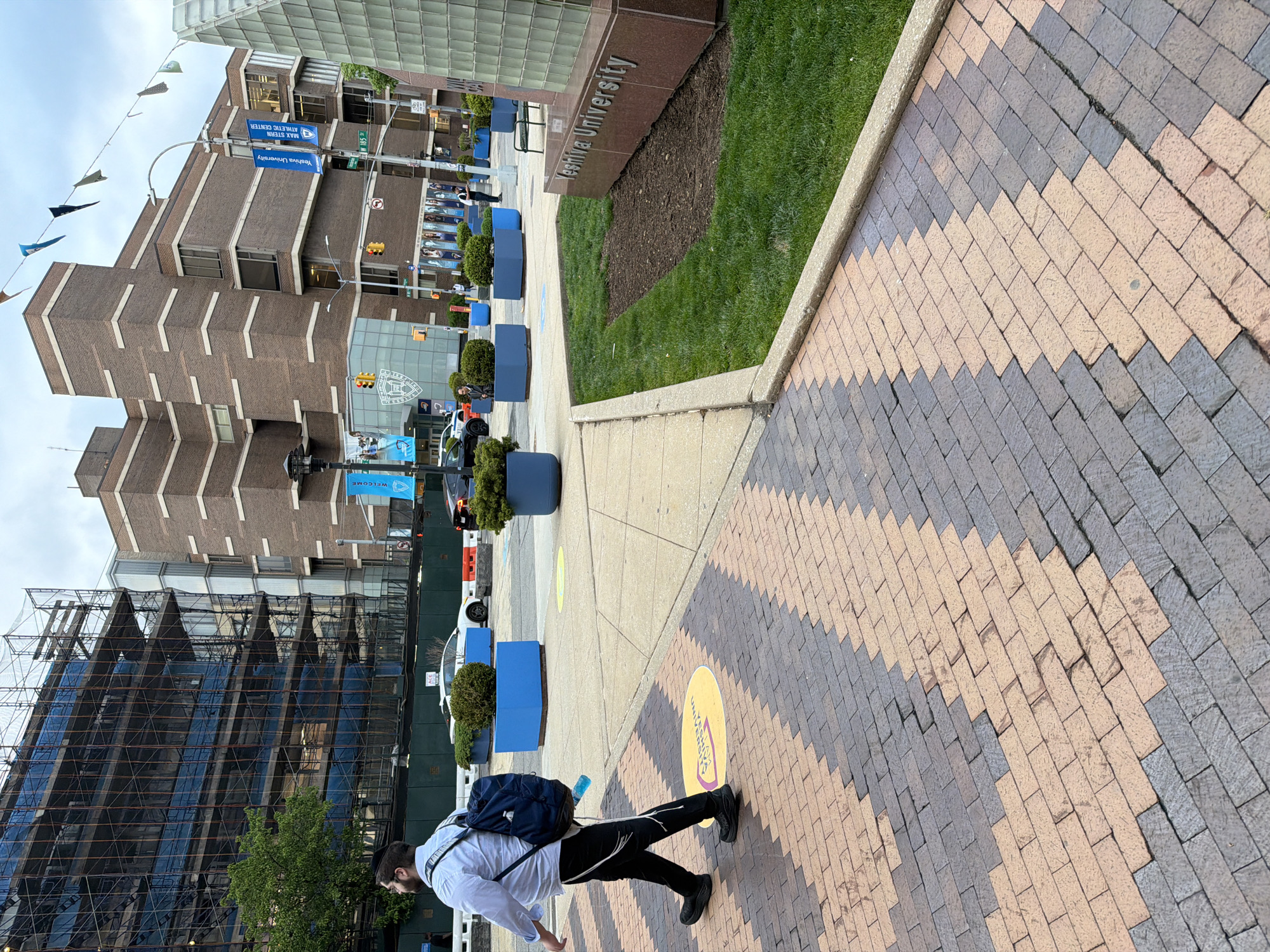 A street scene in New York City, United States of America, features a person walking on a patterned brick sidewalk in front of Yeshiva University. The person, wearing a light-colored shirt, dark pants with a white stripe, and dark shoes, carries a dark backpack and a water bottle. The sidewalk leads towards a street with traffic signals and several blue planters. Buildings, including one under construction with scaffolding and netting, and another brick building with signage indicating Yeshiva University and the Max Stern Athletic Center, line the street. Banners hang between buildings, and a sign bearing the university name is visible near a green space.