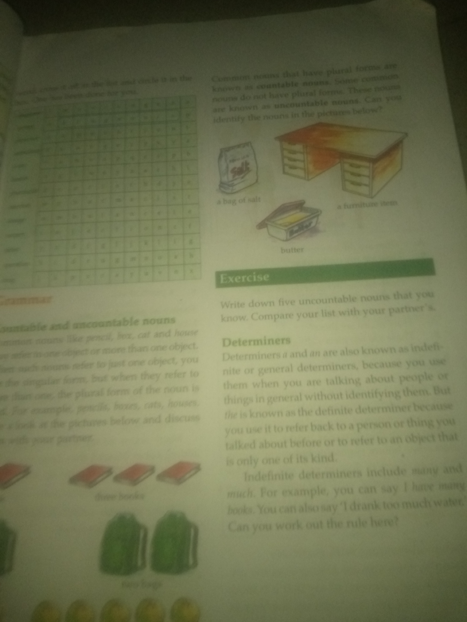 The image is a page from a book, likely a grammar textbook, discussing countable and uncountable nouns. On the left side of the page, there is a word search puzzle grid filled with letters. Below the puzzle, there is a section titled "Grammar" which defines countable and uncountable nouns with examples. Further down, there are illustrations of three books and two backpacks, with captions "three books" and "two bags". On the right side of the page, there are illustrations of a bag of salt, a desk, and a container of butter. Above these illustrations, there is text defining common nouns that have plural forms and those that do not. Below the illustrations, there is a section titled "Exercise" asking to write down five uncountable nouns. Another section discusses "Determiners," specifically indefinite and definite determiners, with examples. The overall image is a scanned page of an educational text. There is no information about Jauro Yinu, Nigeria in this image.