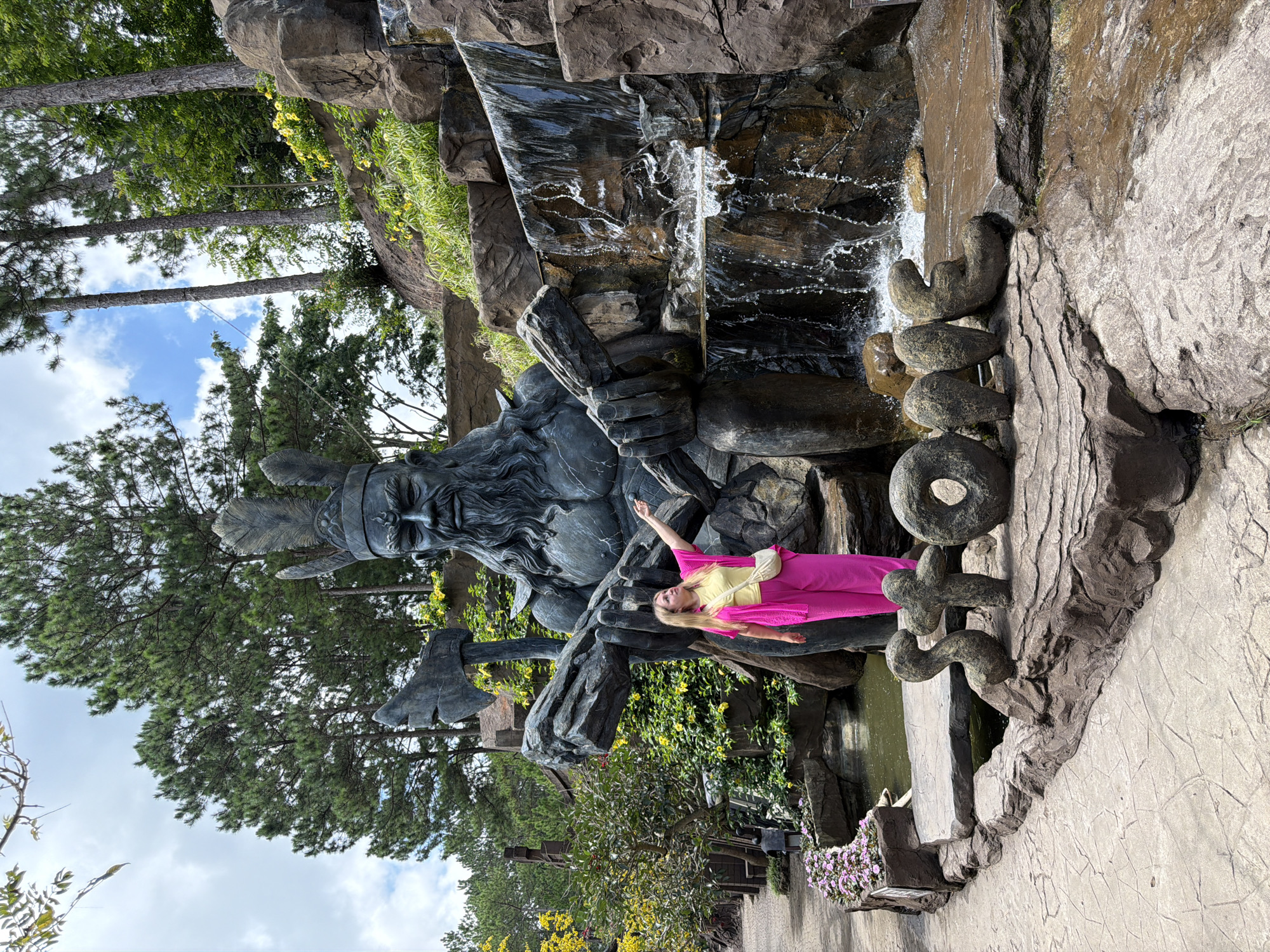 This image depicts a tourist posing in front of a large, imposing statue of a man with a beard, wearing a headdress and holding axes. The statue is situated in a garden-like environment with a waterfall cascading behind it. In the foreground, large stone letters spell out "STONE." A woman, dressed in a bright pink outfit and a yellow top, is standing to the left of the statue, pointing towards it. She is wearing a small crossbody bag. The background features pine trees and lush greenery, suggesting an outdoor natural setting. The overall scene is likely a tourist attraction in Da Lat, Vietnam, known for its natural beauty and various themed gardens and statues.