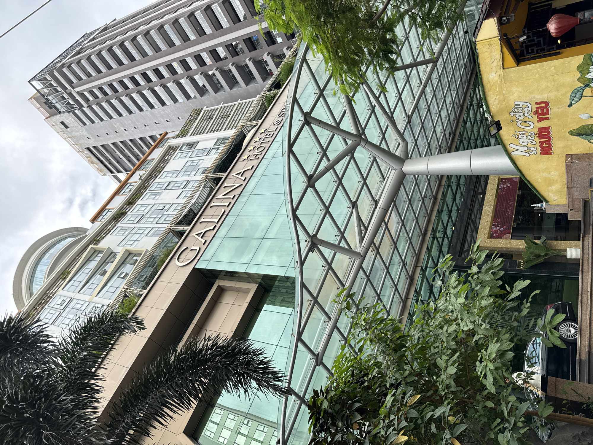 This is a low-angle, eye-level, exterior, full-shot photograph of the Galina Hotel & Spa in Nha Trang, Vietnam. The Galina Hotel & Spa is a multi-story building with a prominent glass facade and a modern, curved glass awning covering the entrance. The hotel's name is visible in large, stylized letters on the facade. To the right of the Galina Hotel & Spa, a taller, more modern, grey skyscraper with balconies rises into the sky. In the foreground, lush green trees partially obscure the view of the hotel entrance. A black sedan is parked in front of the hotel, with its rear facing the camera. A person in a white shirt and black pants is standing next to the car. To the far right, a yellow building with Vietnamese writing and a red lantern is visible. The sky is overcast.