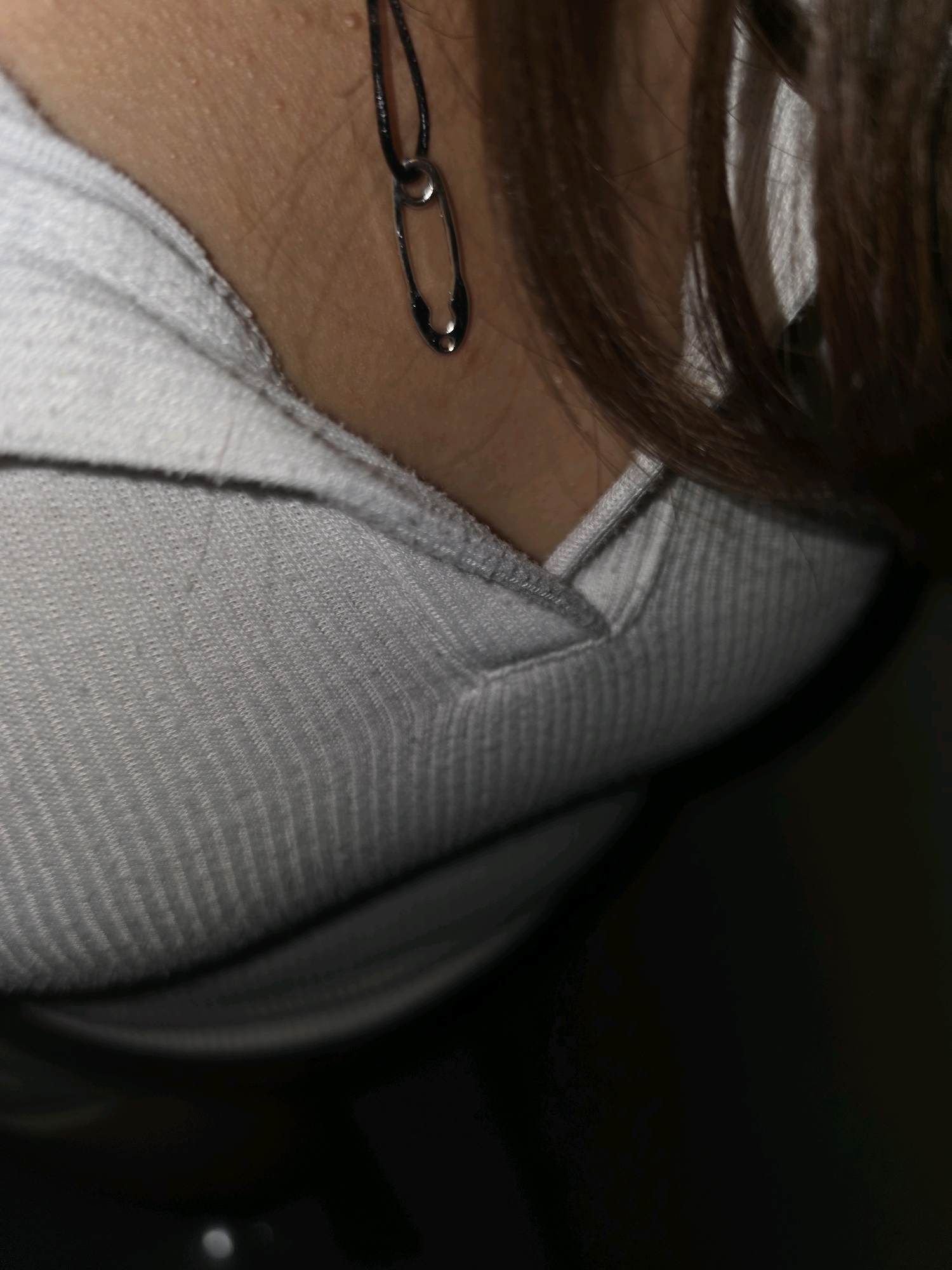 The image shows a close-up of a person's neck, wearing a grey ribbed shirt and a safety pin earring. The background is dark and out of focus. The location is not specified in the image, and therefore cannot be determined to be in Pintomakmur, Indonesia.