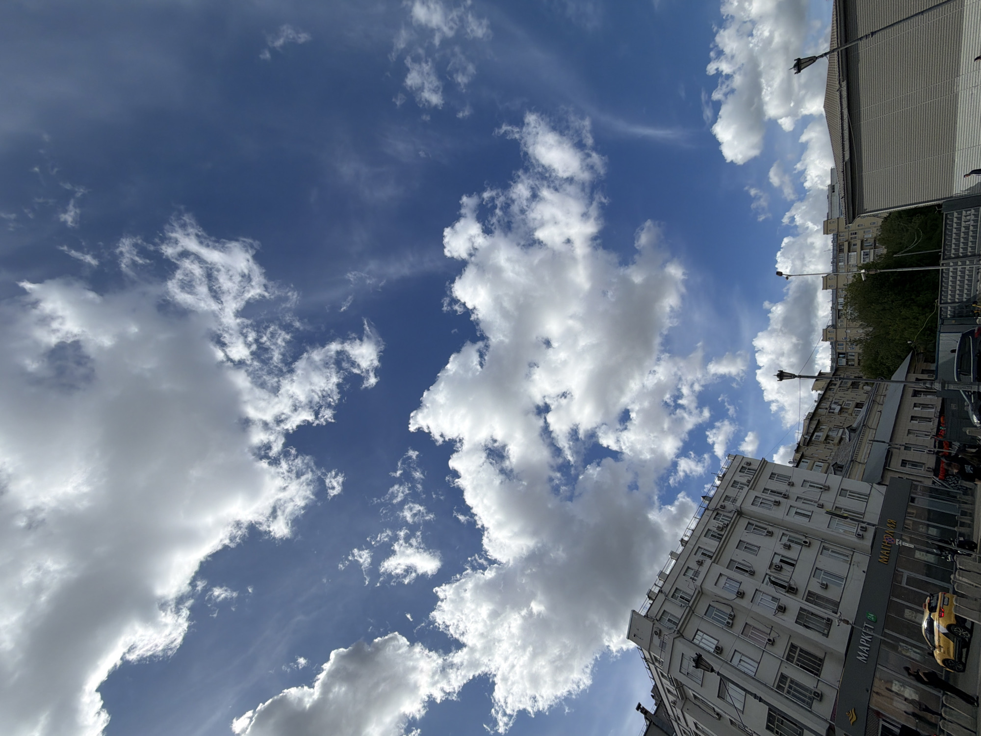 The image shows a view of a city street in Unknown, Unknown, angled upward to capture a bright blue sky filled with fluffy white cumulus clouds. On the right side of the frame, several multi-story buildings line the street. The buildings are primarily light-colored with numerous windows and what appear to be air conditioning units. A storefront is visible on the lower floors of one building, with the signage "MAPKET 24" written in a script that includes Cyrillic characters, above which is "МАРКЕТ". Below this is an unknown language.
A few figures are visible near the base of the building, along with parked vehicles. Above the buildings, treetops and street lights can be seen.