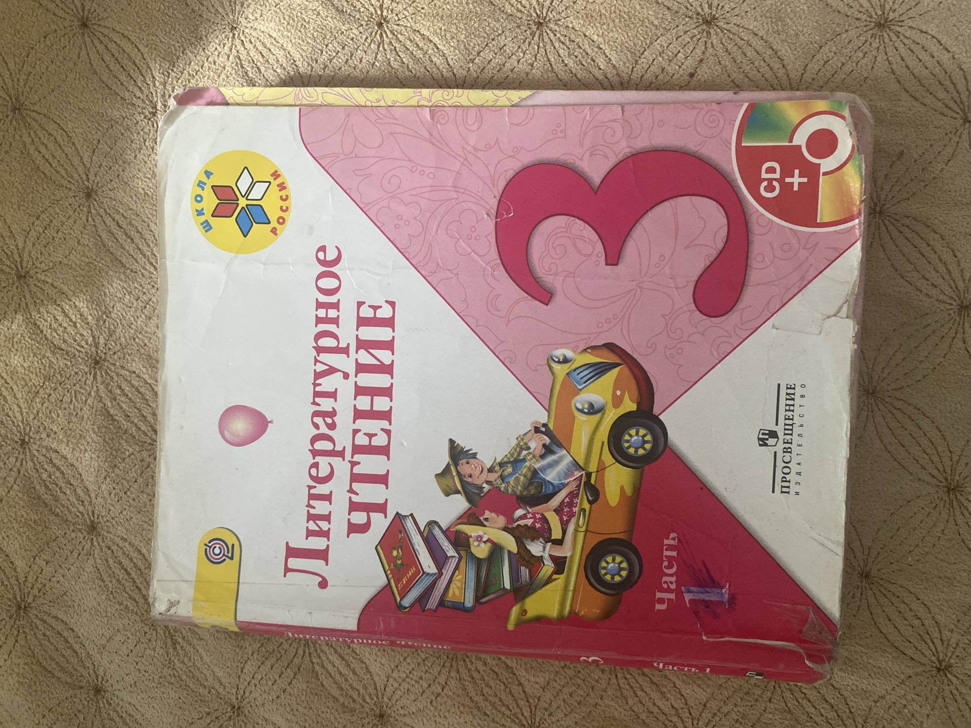 The image shows the cover of a Russian language textbook for children, titled "Литературное Чтение" (Literary Reading), specifically part 3. The cover features colorful illustrations, including a stylized car with two children in it, a stack of books, and a balloon. There are also logos indicating a "School of Russia" program and a CD-ROM. The textbook is lying on a textured, tan-colored surface, likely a carpet or blanket. There are no discernible landmarks or elements in the image that can be used to determine its location in the city of Kizilyurt, Russia. The focus is solely on the book itself.