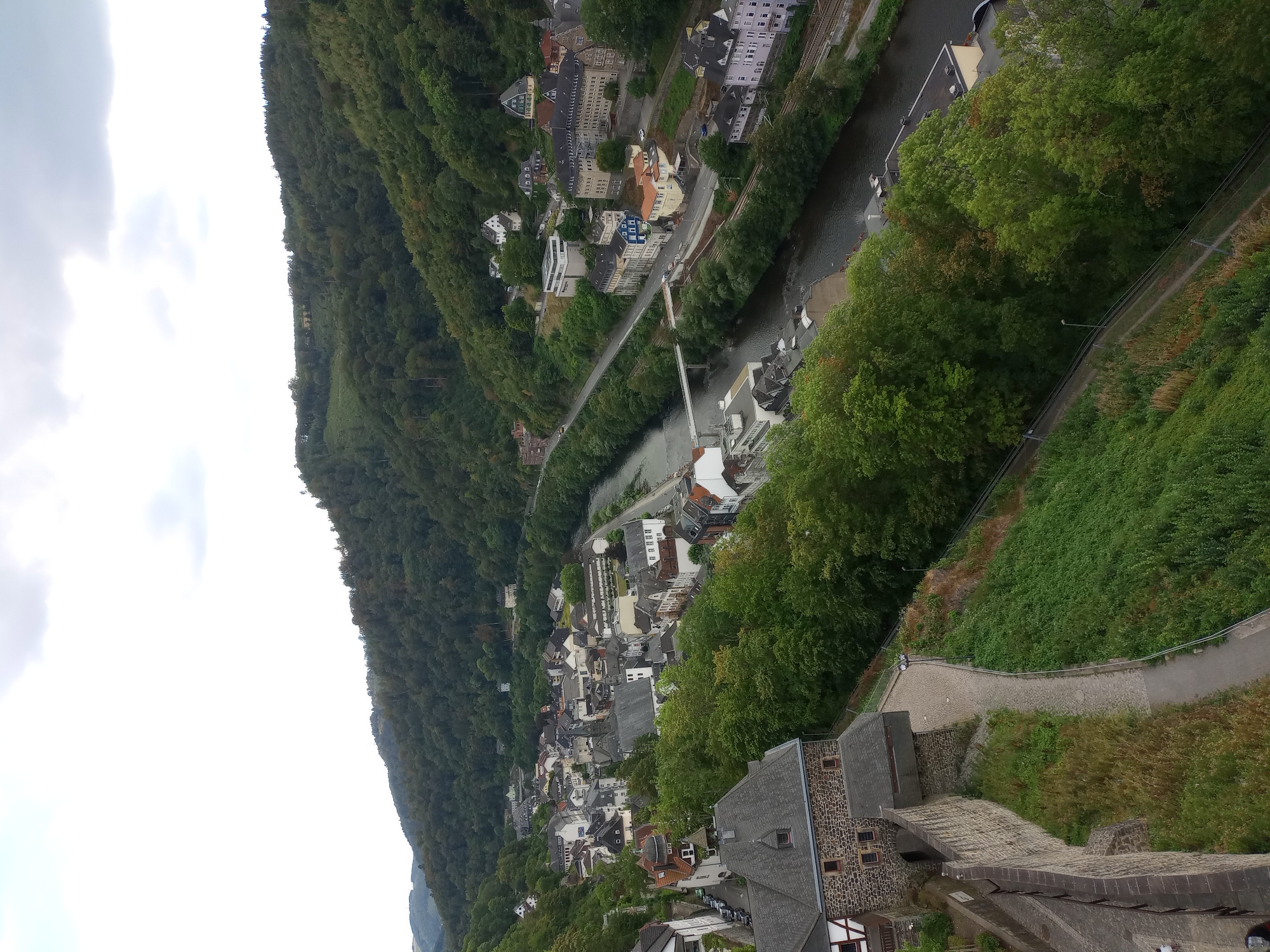 This is a high-angle, wide shot of the town of Altena, Germany, nestled within a lush, forested valley. A dark, winding river bisects the town. Buildings, primarily residential and commercial with pitched roofs and varying architectural styles, are densely packed along the riverbanks and the hillsides. A road runs parallel to the river, with a bridge crossing it. Further up the hill, more houses and buildings are visible, interspersed with trees. A stone wall, part of what appears to be a castle or fortification, occupies the left foreground. A paved path follows the wall along the hillside. The sky is overcast with scattered clouds. There are no discernible people or vehicles in the immediate foreground or midground. The overall scene depicts a historic German town integrated with its natural environment.