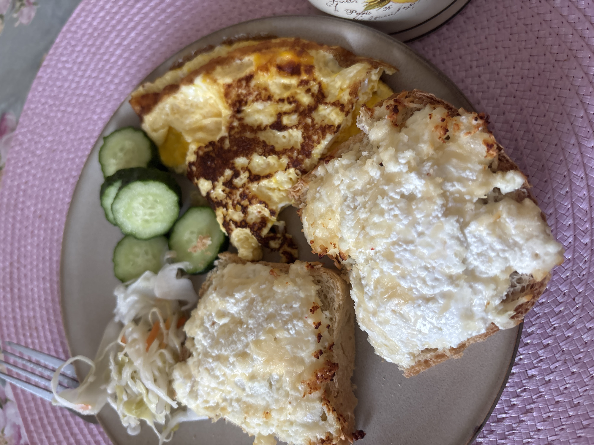 A plate of breakfast food is presented on a pink woven placemat. The plate contains two slices of toasted bread topped with a white, creamy cheese spread, an omelet with browned edges, sliced cucumbers, and a small portion of coleslaw. A fork is visible in the upper left corner, partially resting on the coleslaw. The background is softly blurred, suggesting an indoor setting. The lighting appears to be natural, coming from above and to the left. There are no individuals or specific landmarks of Naberezhnye Chelny, Russia visible in the image.
