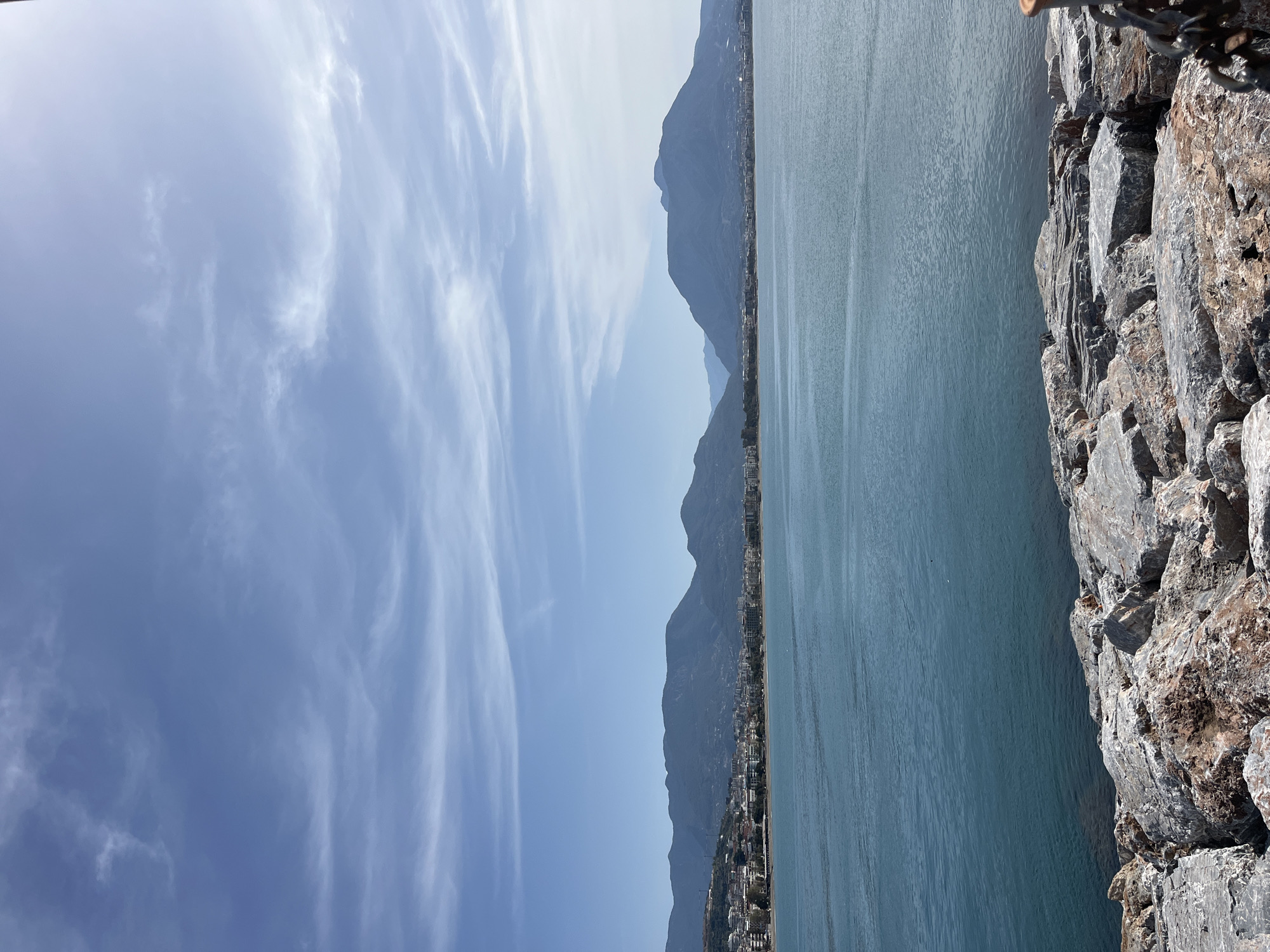 The image captures a coastal scene in Alanya, Turkey, featuring a view of the Mediterranean Sea from a rocky edge. In the foreground, a rough, gray rock barrier forms the right edge of the image, extending upwards. The calm, blue water of the Mediterranean stretches out towards the horizon, occupying a significant portion of the scene. In the distance, the rugged Taurus Mountains are visible, partially obscured by a light haze, and a coastal city, Alanya, stretches along the shoreline, nestled at the foot of the mountains. The sky is predominantly blue with streaks of cirrus clouds that extend diagonally across the frame. The light suggests it is a daytime shot with clear weather.