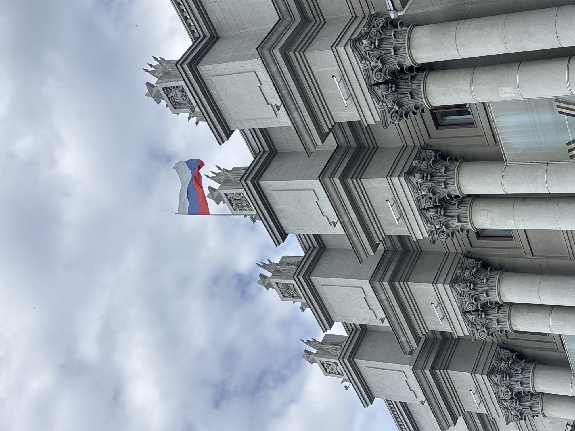 This is a photograph of the Novokuybyshevsk Oil Refinery Administration Building, located in Novokuybyshevsk, Russia. The image is taken from a low angle, looking up at the facade of the building. The building is made of concrete and features classical architectural elements such as columns with ornate capitals and decorative sculptural details on the upper sections. A Russian flag is prominently displayed on a flagpole to the left of the frame. The sky is overcast with clouds. There are no people visible in the image.