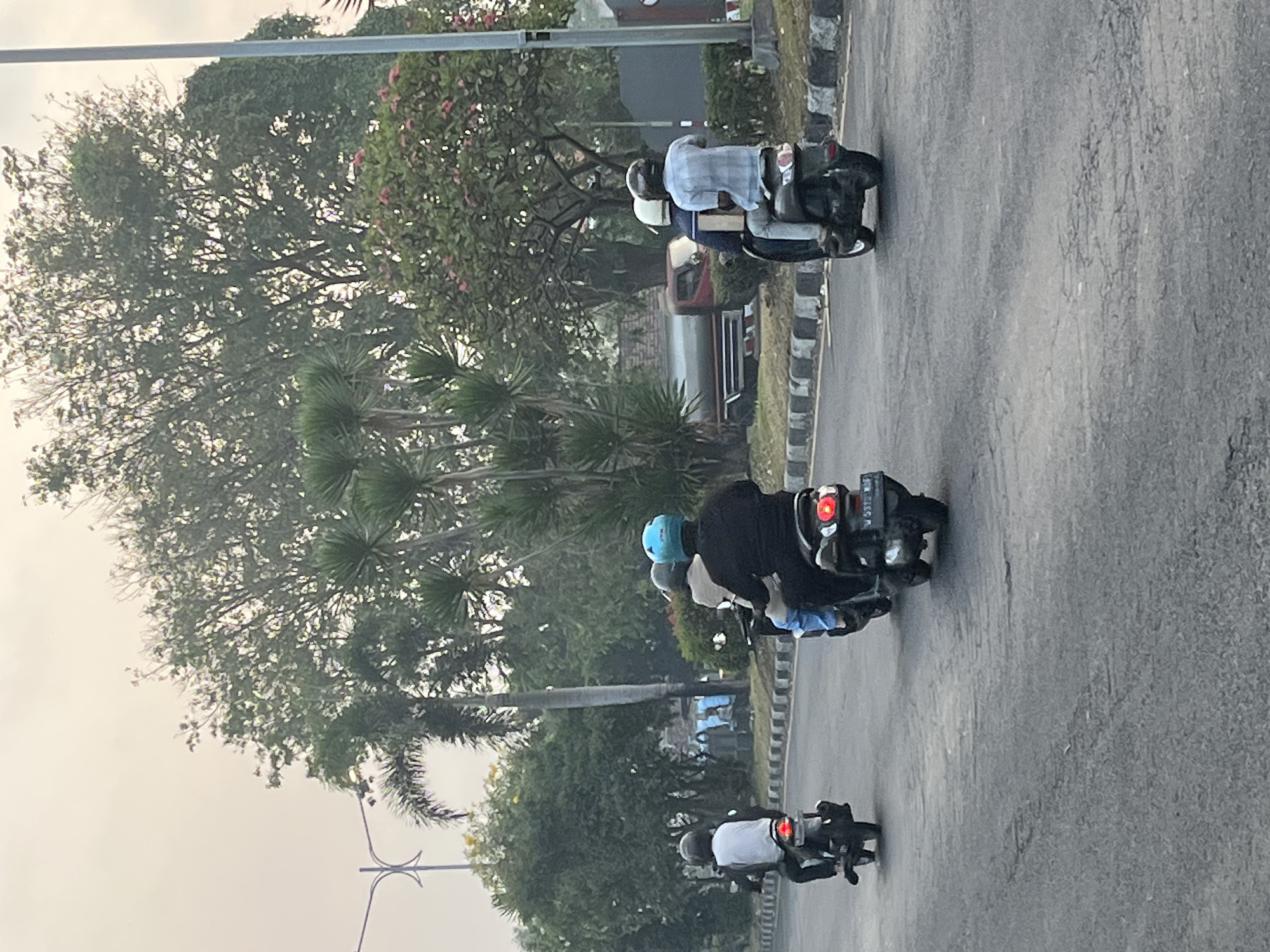 This is a photograph of a road in Kludan, Indonesia. Four individuals are riding motorcycles. The person on the leftmost motorcycle is wearing a white shirt and a dark helmet. The person behind them is wearing a light blue helmet and a dark jacket. The next motorcycle has two passengers. The rider is wearing a dark helmet and a dark jacket, and the passenger is wearing a light blue helmet. The farthest motorcycle has two passengers, both wearing helmets. The rider is wearing a light blue plaid shirt and the passenger is wearing a white shirt. The road is paved and appears to be in good condition, with a curb on the right side. Trees and foliage are visible on either side of the road, and a utility pole is on the right. The sky is overcast.