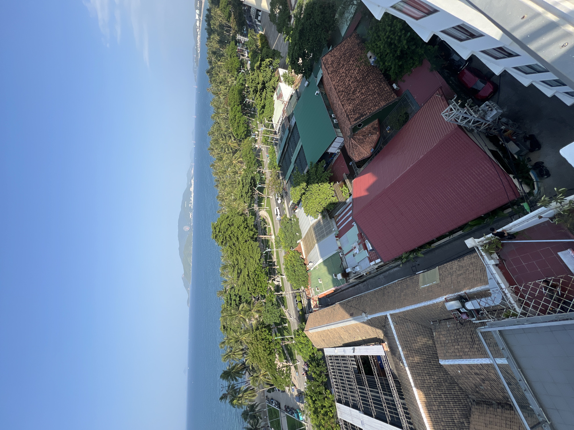 An aerial, wide-angle, eye-level shot captures a panoramic view of Nha Trang, Vietnam. The foreground is dominated by the rooftops of various buildings, characterized by their tiled and metal coverings, and interspersed with lush green trees. A clear blue sky stretches across the upper half of the image, meeting the tranquil blue ocean at the horizon. Beyond the immediate rooftops, a tree-lined boulevard separates the urban landscape from the picturesque coastline. Palm trees and other dense foliage form a vibrant green belt along the beach, with the ocean waves gently lapping the shore. In the distance, a mountainous landscape provides a scenic backdrop, and a Ferris wheel is visible on one of the hills. The presence of boats on the water suggests a bustling maritime activity. The scene is bathed in the soft light of what appears to be late afternoon or early morning.