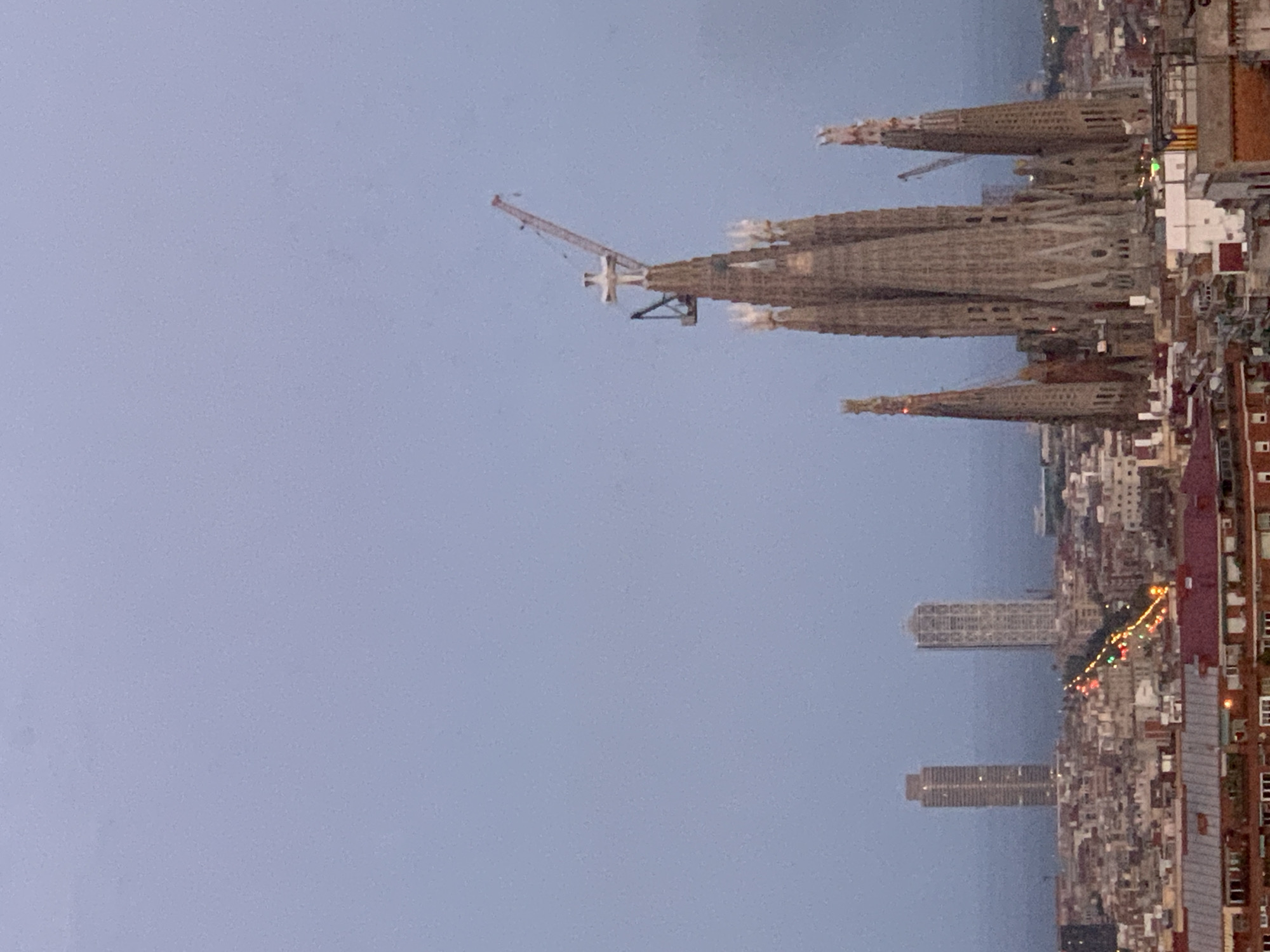 The image captures a panoramic view of Barcelona, Spain, dominated by the iconic Sagrada Familia basilica, which is clearly under construction with a crane visible at its peak. The basilica's intricate Gothic architecture and numerous spires are prominent. In the background, two modern skyscrapers stand tall against a hazy sky, with a city skyline composed of numerous buildings filling the midground. A street with glowing car lights suggests the setting is either dawn or dusk. The overall scene depicts a blend of historical and contemporary architecture within a bustling urban environment.