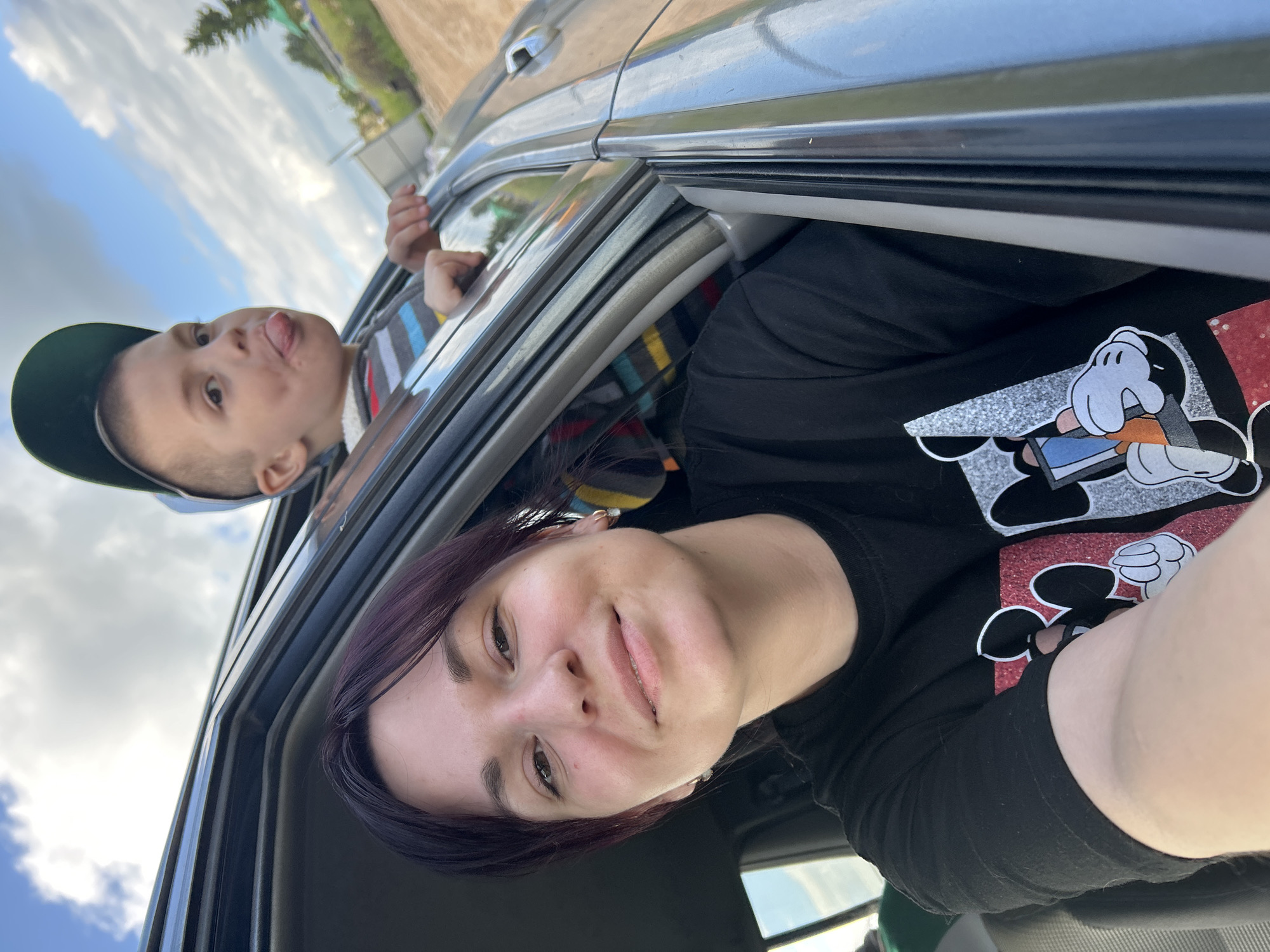 In the city of Fokino, Russia, a selfie is taken from inside a car. A woman with dark hair and bangs, wearing a black t-shirt with Mickey Mouse graphics, smiles at the camera. Her arm is extended, capturing the selfie. To her right, a young boy with short hair wears a green cap and a striped shirt. He sticks his tongue out of the open car window, looking towards the camera. The car's interior is visible, with gray trim and a black seat. Outside the car, a cloudy sky with blue patches is visible, along with greenery and trees, suggesting a rural or suburban setting. The overall scene depicts a casual, outdoor moment shared between a woman and a child during a car ride.
