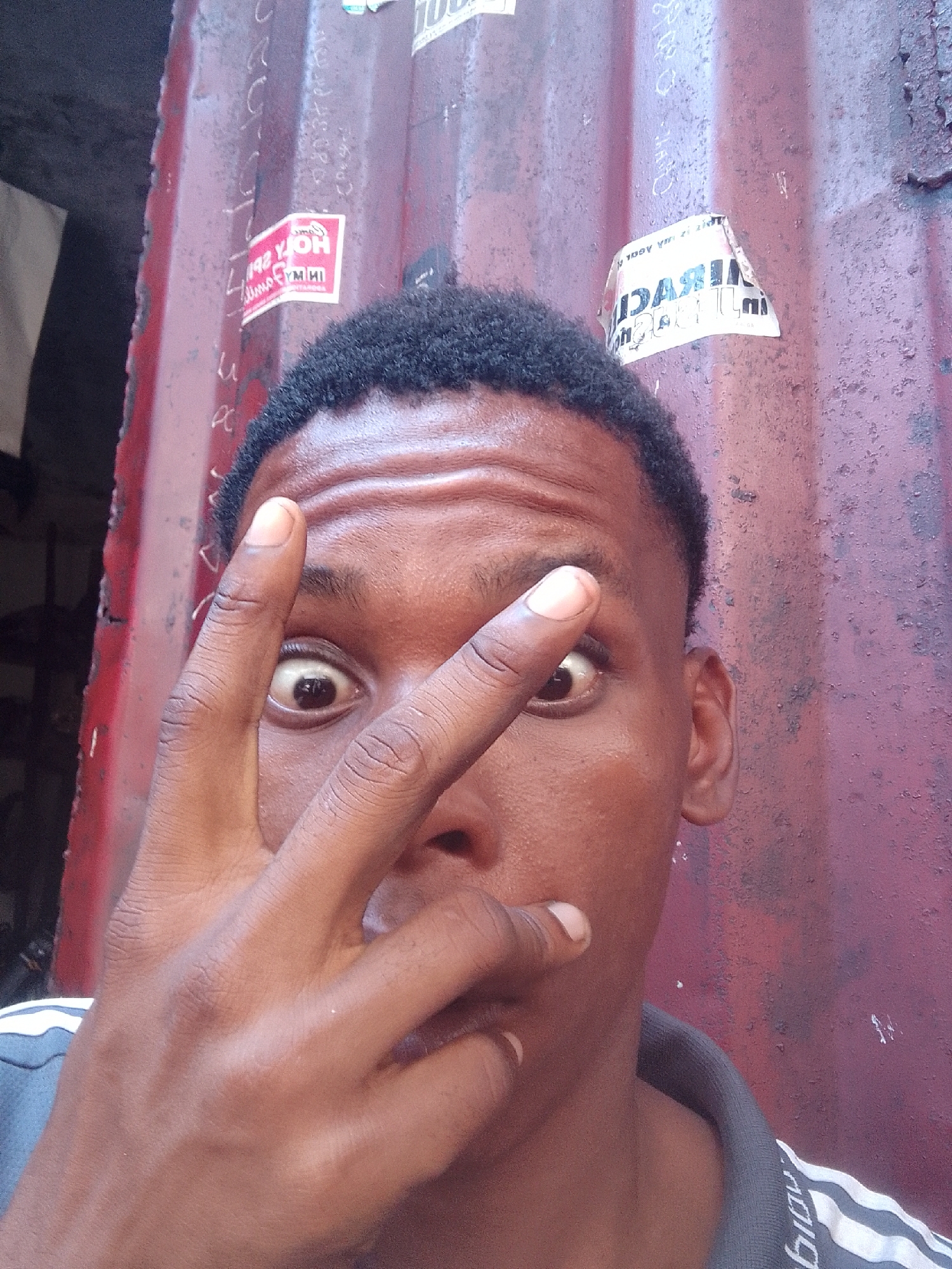 A medium shot captures a person of African descent with short, dark hair, partially obscuring their face with their hand in a "peace" sign. Their eyes are wide and expressive. The individual is wearing a shirt with white stripes on the shoulder.
The background consists of a corrugated metal wall, peeling and stained, with remnants of stickers or posters affixed to it. The setting is identified as Onitsha, Nigeria. The image appears to be a candid snapshot taken in an urban environment.