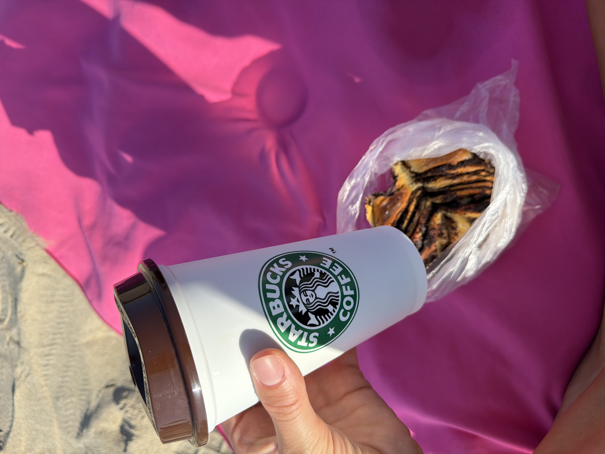 In Urbanització Cales I Talaies, Spain, a person's hand, with neatly manicured fingernails, holds a white Starbucks coffee cup. The cup features the iconic green Starbucks logo. The cup is positioned over a plastic bag containing a slice of chocolate babka. The scene is set against a bright pink fabric, likely a beach towel or blanket, with sand visible in the upper left corner. Shadows, cast by an unseen object or person, create a dramatic effect on the pink fabric. The lighting suggests it is a sunny day, likely outdoors.