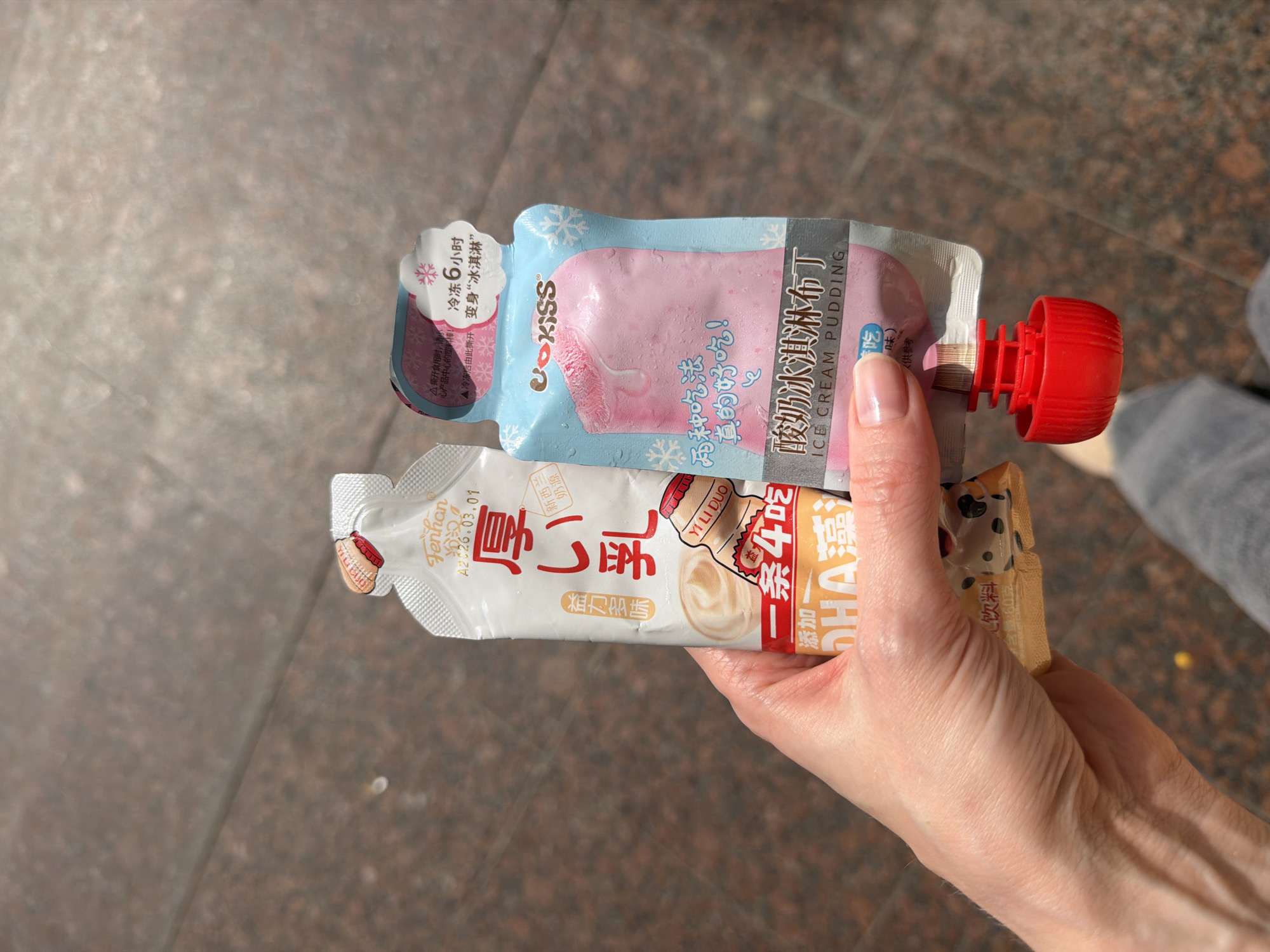 A person's hand holds two food pouches against a blurred gray outdoor background, possibly a city sidewalk. The upper pouch is light blue and pink with the brand "UOKISS" and text indicating it's ice cream pudding, with a red plastic nozzle. The lower pouch is white with red and brown accents, featuring the brand "YI LI DUO" and Chinese characters, along with text about "DHA" and "Fenhan." The light suggests an outdoor setting, though the precise location (Moscow, Russia) cannot be verified from the visual data.