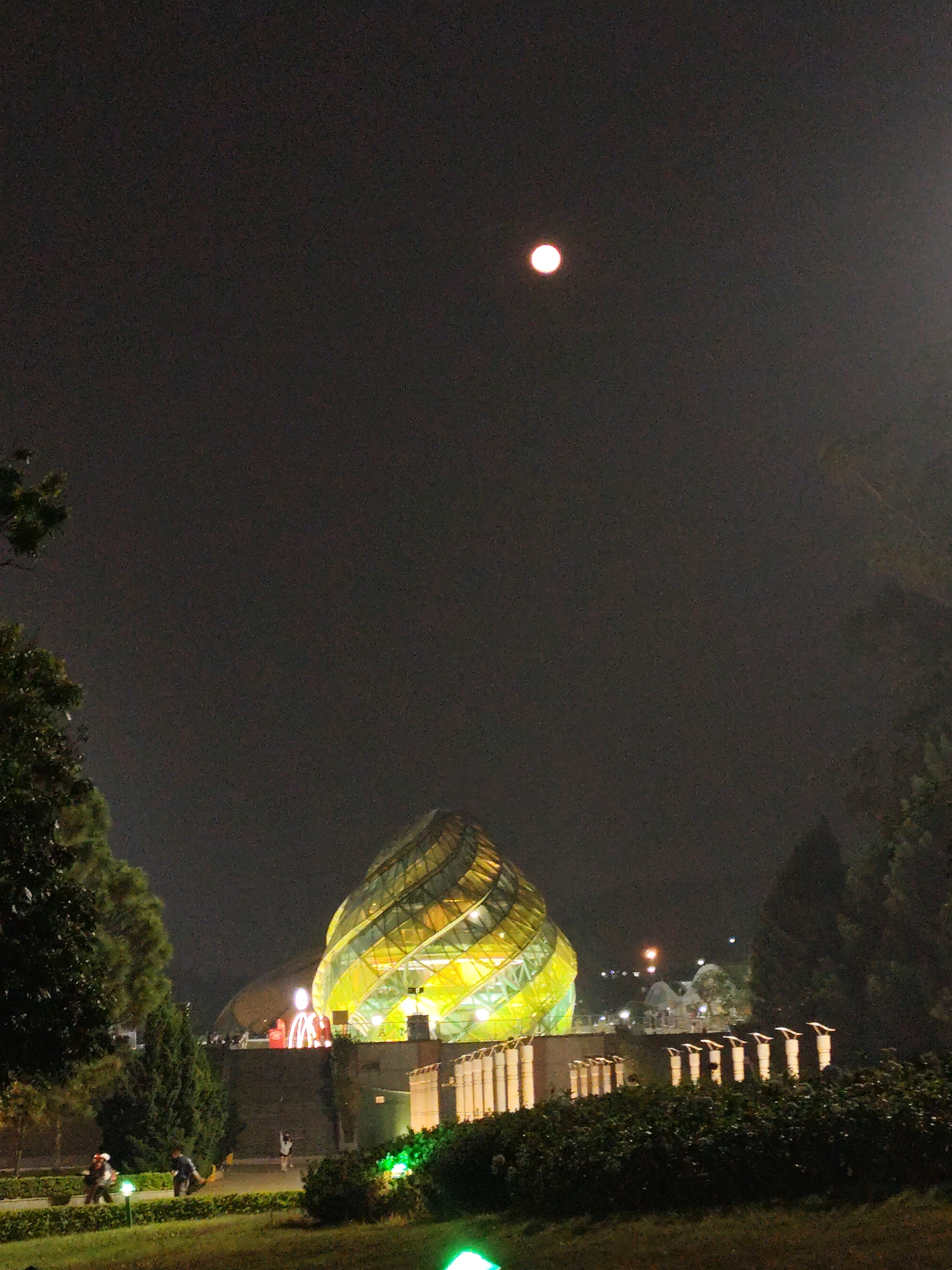 At night, the image shows the Da Lat Cathedral, a building in Da Lat, Vietnam. The structure has a yellow and green glass exterior in a spiraling shape. It is illuminated, contrasting with the dark sky above. A full moon shines brightly above and behind the cathedral. In the foreground, manicured lawns and greenery are visible, with small, lighted posts lining the garden path. People are walking along the path. In the background, other lights suggest the presence of city structures.
