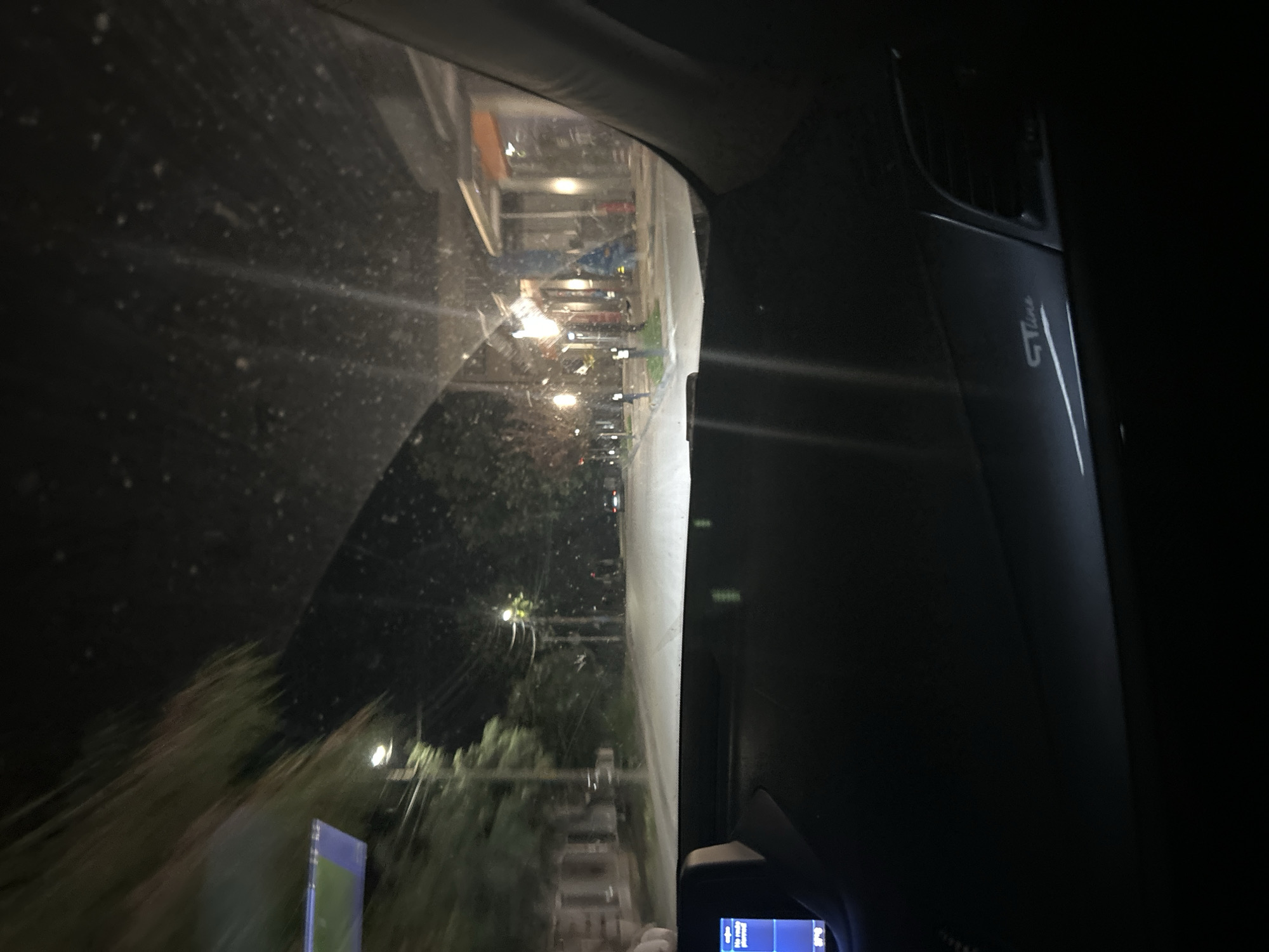 The image is a nighttime or low-light photograph taken from inside a vehicle, looking out through the windshield and side window. The view is angled, as if the camera is tilted or the vehicle is on an incline.
The scene outside appears to be a street or an area with some buildings and trees. Lights from street lamps and building interiors are visible, casting reflections on the vehicle's windows, which are speckled with dirt or moisture. Some foliage is visible in the foreground, blurred due to motion. In the distance, streetlights line what looks like a road. A blue and green sign is partially visible on the left side of the frame.
Inside the vehicle, the interior is dark. A portion of the car's dashboard is visible on the right, including what appears to be an air vent and a silver trim piece with the text "GT Line" embossed on it. At the bottom right, a small screen with some blue and white text is visible, possibly a car display or a phone screen.
The overall impression is one of being in a car traveling through a semi-urban or suburban area at night. There are no clear indications of specific activities or events taking place beyond the general context of a drive. The low light conditions suggest it is either nighttime or dusk/dawn. There are no discernible emotions conveyed by any subjects, as people are not clearly visible.