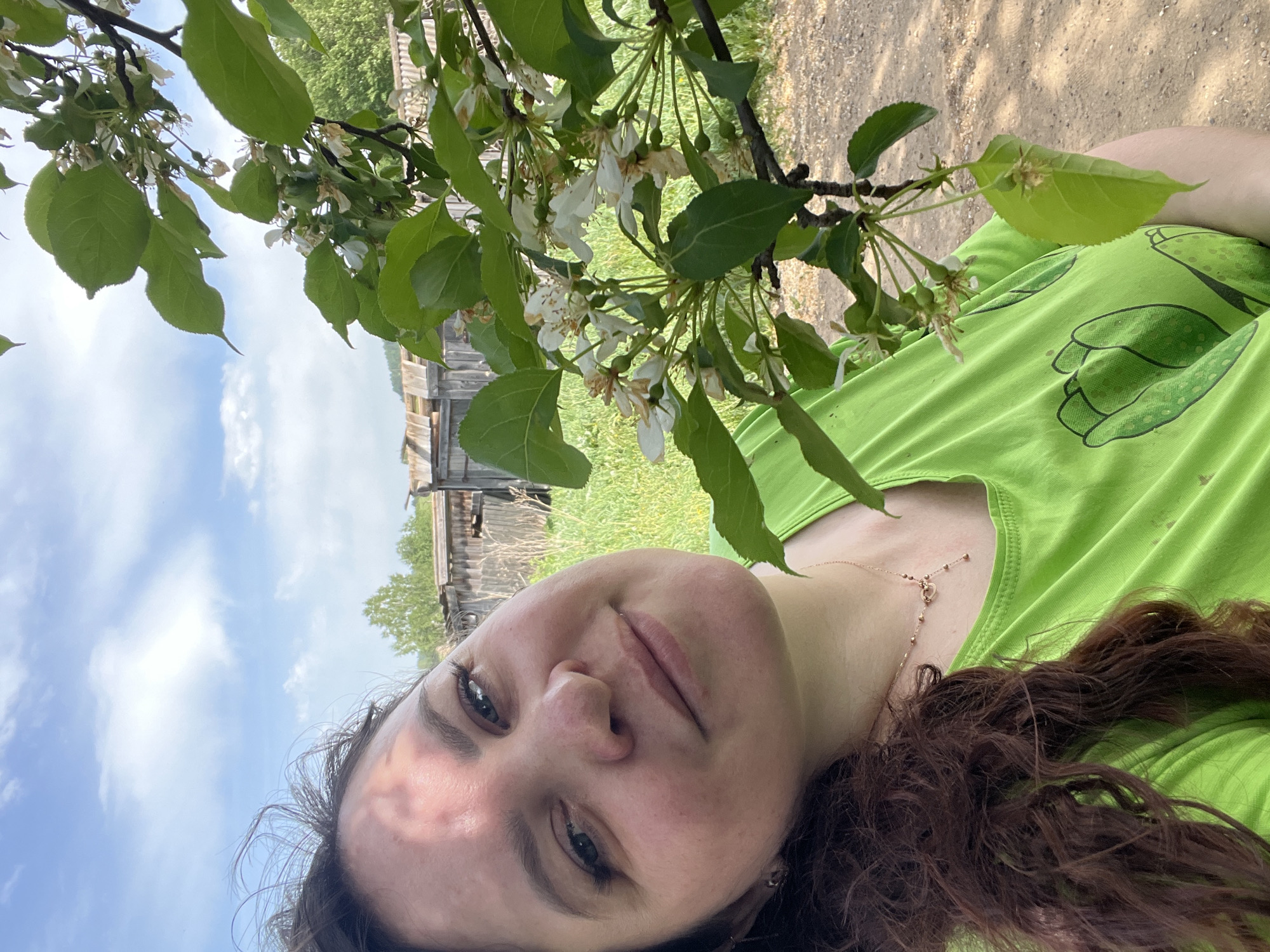 A woman with brown curly hair and fair skin is taking a selfie outdoors. She is wearing a light green t-shirt and a gold necklace. Above her head, a tree branch with green leaves and white blossoms partially obscures the sky, which has a few scattered clouds. In the background is a rustic wooden building, possibly a barn or shed, and a grassy field. A dirt path can be seen at the upper right corner of the frame. The location is likely in or near the city of Panteleevka, Russia.