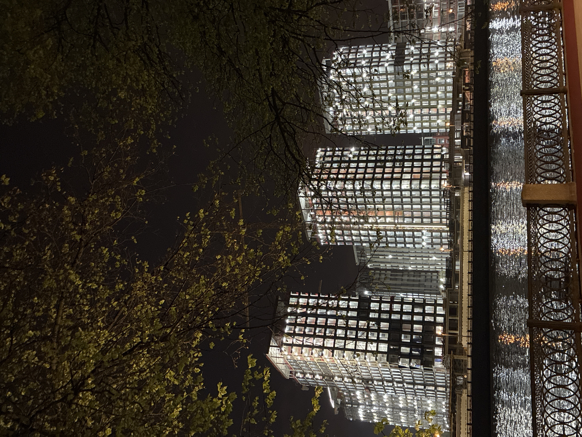 The image shows a nighttime scene in Moscow, Russia, with three tall buildings reflected in the water. The buildings have a grid-like appearance due to numerous illuminated windows. On the right side, a decorative metal railing is visible, and on the left, trees with green leaves are partially obscuring the view. The water reflects the lights of the buildings, creating a shimmering effect.