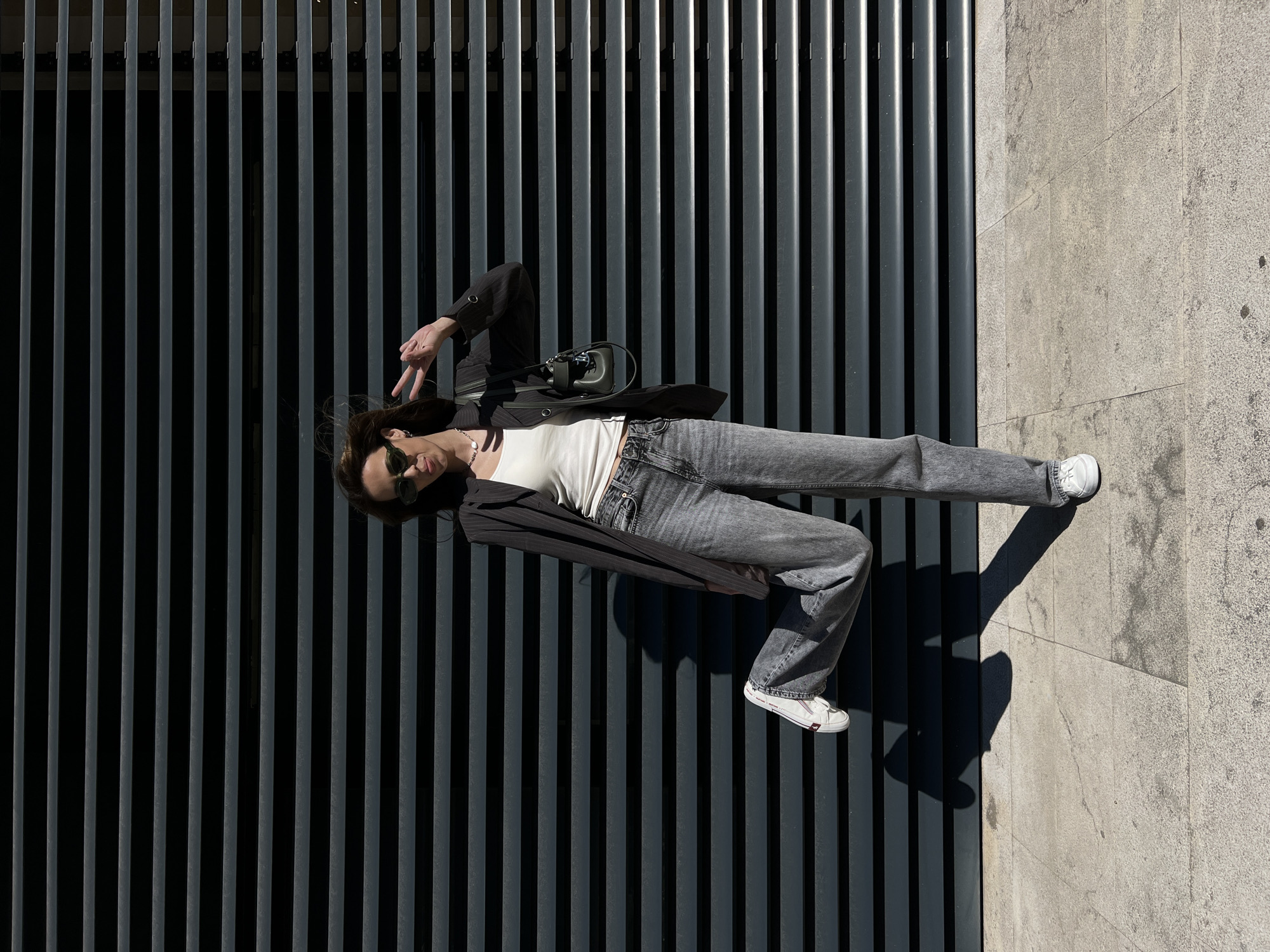 A woman is posing for a photograph against a slatted wall in Bar, Montenegro. The woman is wearing a black blazer, a white tank top, grey jeans, and white sneakers. She has sunglasses on. Her head is near the upper left portion of the frame, and her feet are near the lower right. The slatted wall is dark grey, and a light grey sidewalk is to the right of the wall. The slats run vertically and fill most of the image. The woman is lying down on the wall, with her body angled slightly. She is making a peace sign with her left hand. A purse rests on her torso.
