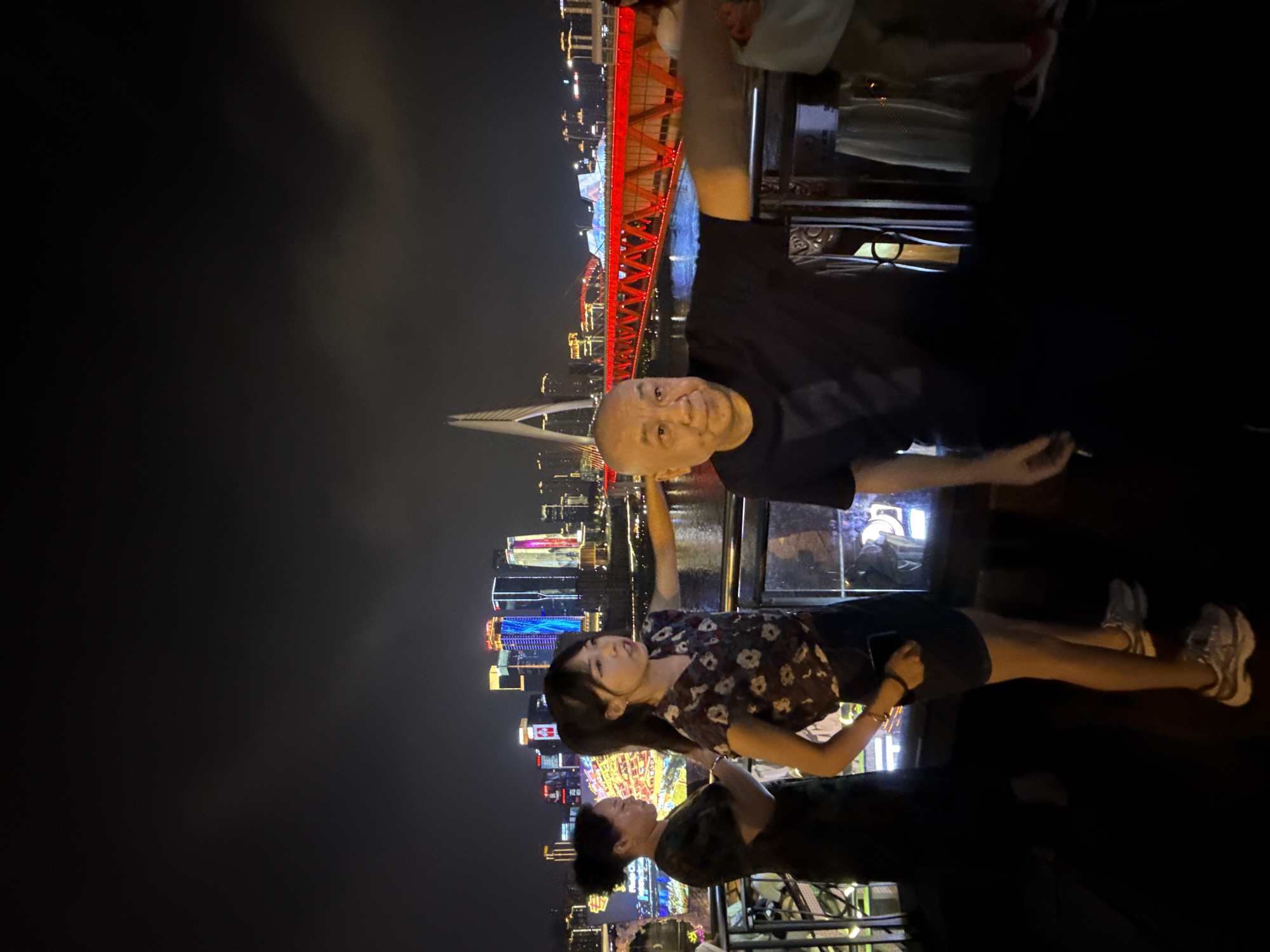 A group of people stand on a platform at night, overlooking a city skyline with illuminated buildings and a brightly lit red bridge. In the foreground, an older man with a bald head wears a black t-shirt and points his right arm towards the right of the frame. To his left, a young woman with dark hair in a ponytail wears a floral print blouse and a dark skirt, holding a phone in her left hand. Behind her, another woman in a dark dress is partially visible. The background features a river and a modern suspension bridge with illuminated cables. The city is identified as being in the Yuzhong District of Chongqing, China.