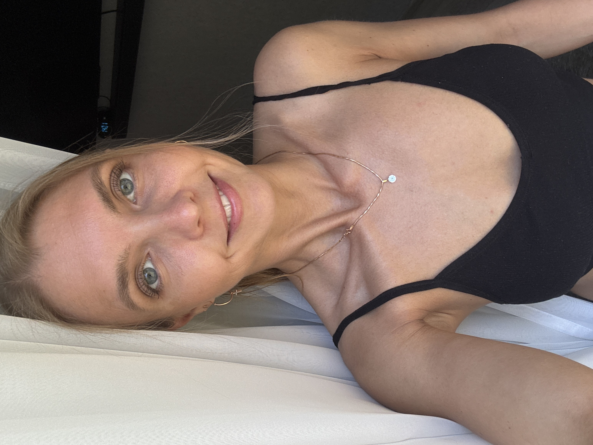 A woman with blonde hair and blue eyes is lying on a white surface, possibly a bed, wearing a black tank top and a necklace with a small pendant. She is smiling at the camera. The background is dark, with what appears to be a television and wall visible. There is no indication of the location being in Chaikovsky, Russia.