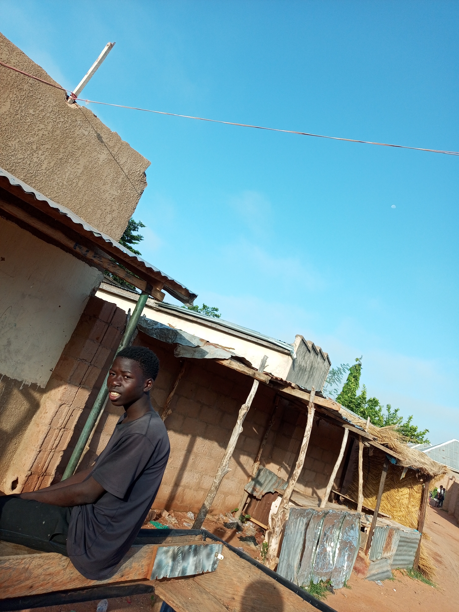A young Black male wearing a dark t-shirt and dark pants is seated on a wooden structure in a dusty outdoor environment. He is looking towards the camera with a slight smile. Behind him, there are several structures made of brick, wood, and corrugated metal. Some structures have thatched roofs. The overall impression is of a rural or semi-urban setting, possibly in a village or the outskirts of a city. The sky is a clear, bright blue with a faint appearance of the moon. The lighting suggests it is daytime. The location is identified as Tempure, Nigeria.