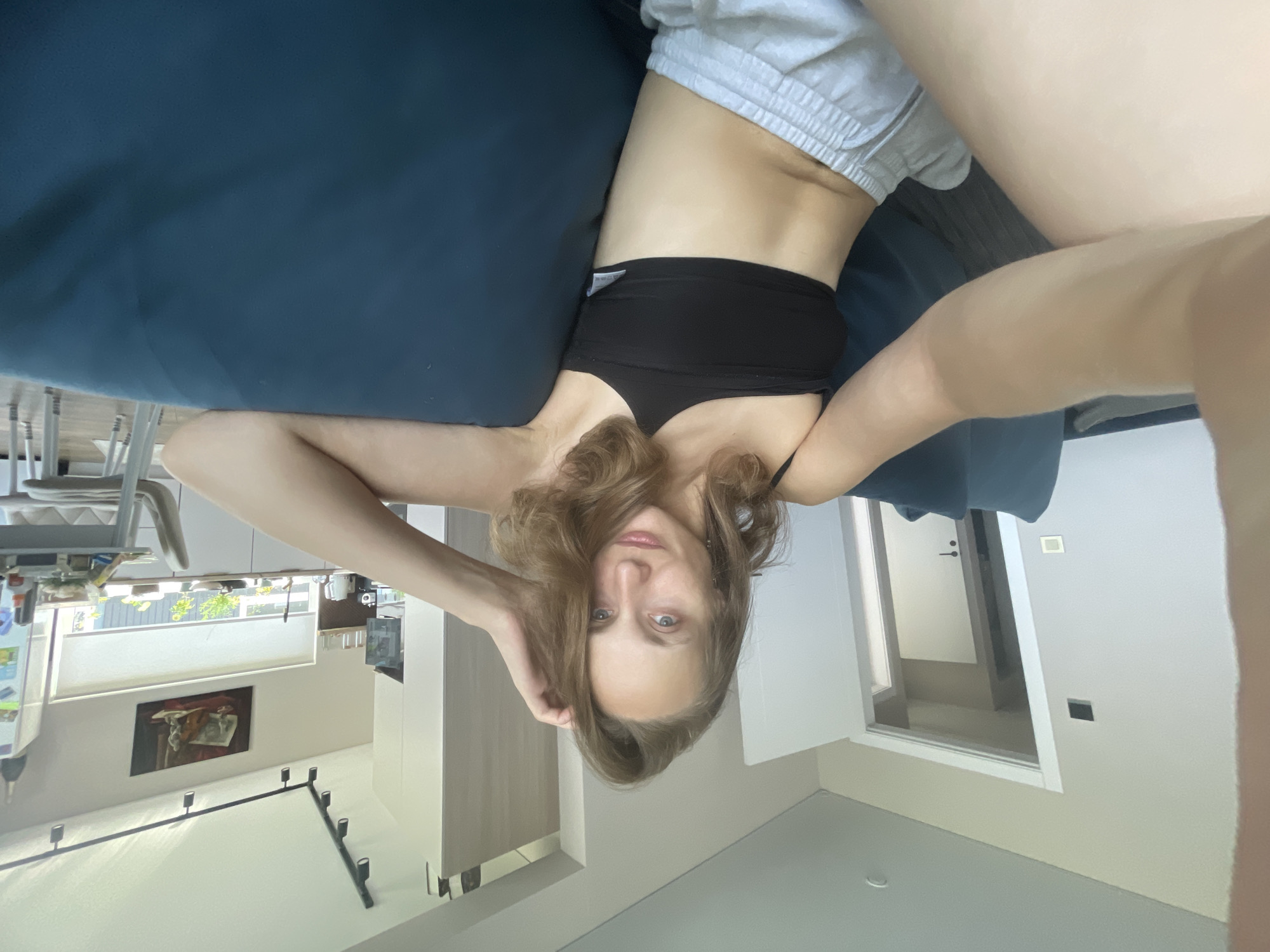 A woman is taking a selfie while sitting on a blue sofa in a modern apartment. She is wearing a black one-shoulder crop top and grey shorts. Her right hand is raised to her head, touching her hair, and her left arm is resting on the back of the sofa. The background shows a living room area with an open doorway to another room, a kitchen with white cabinets and a window, and a painting on the wall. The lighting suggests it is daytime. The specific location in Podshivalovo, Russia, is not discernible from the image.