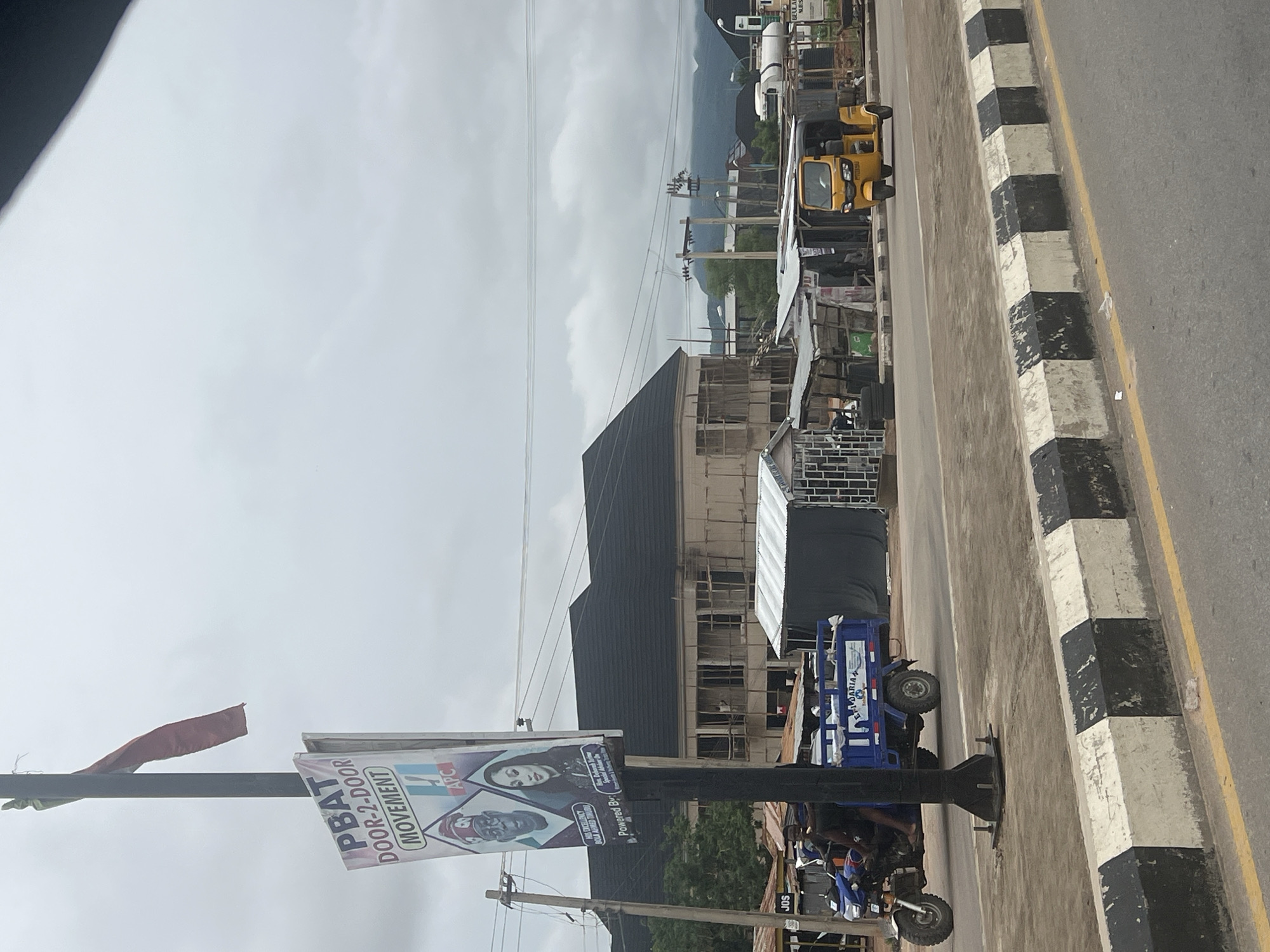 This is a street-level photograph taken on a cloudy day in Jalingo, Nigeria. In the foreground, a black and white striped concrete barrier runs along the edge of the road. To the left of the barrier is a paved road with yellow lane markings.
Dominating the left side of the image is a tall pole with a sign that reads "PBAT DOOR-2-DOOR MOVEMENT". The sign features portraits of two individuals, one older man in a cap and another woman. Below these portraits, text reads "HIS EXCELLENCY BOLA AHMED TINUBU" and "Hon. Deborah Uche Special Adviser On Women & Social Investment". At the bottom of the sign, it says "Powered By". A red flag is tied to the pole above the sign.
Further down the road, a blue motorized tricycle, or "keke," is visible, carrying passengers and goods. To its right, a yellow keke is driving on the road. In the background, there are several buildings, some appearing to be under construction with exposed scaffolding. There are also makeshift structures and market stalls along the roadside. In the far distance, hills or mountains are visible under the overcast sky. The overall scene depicts a typical urban streetscape in Nigeria, with vibrant public transportation and commercial activity.