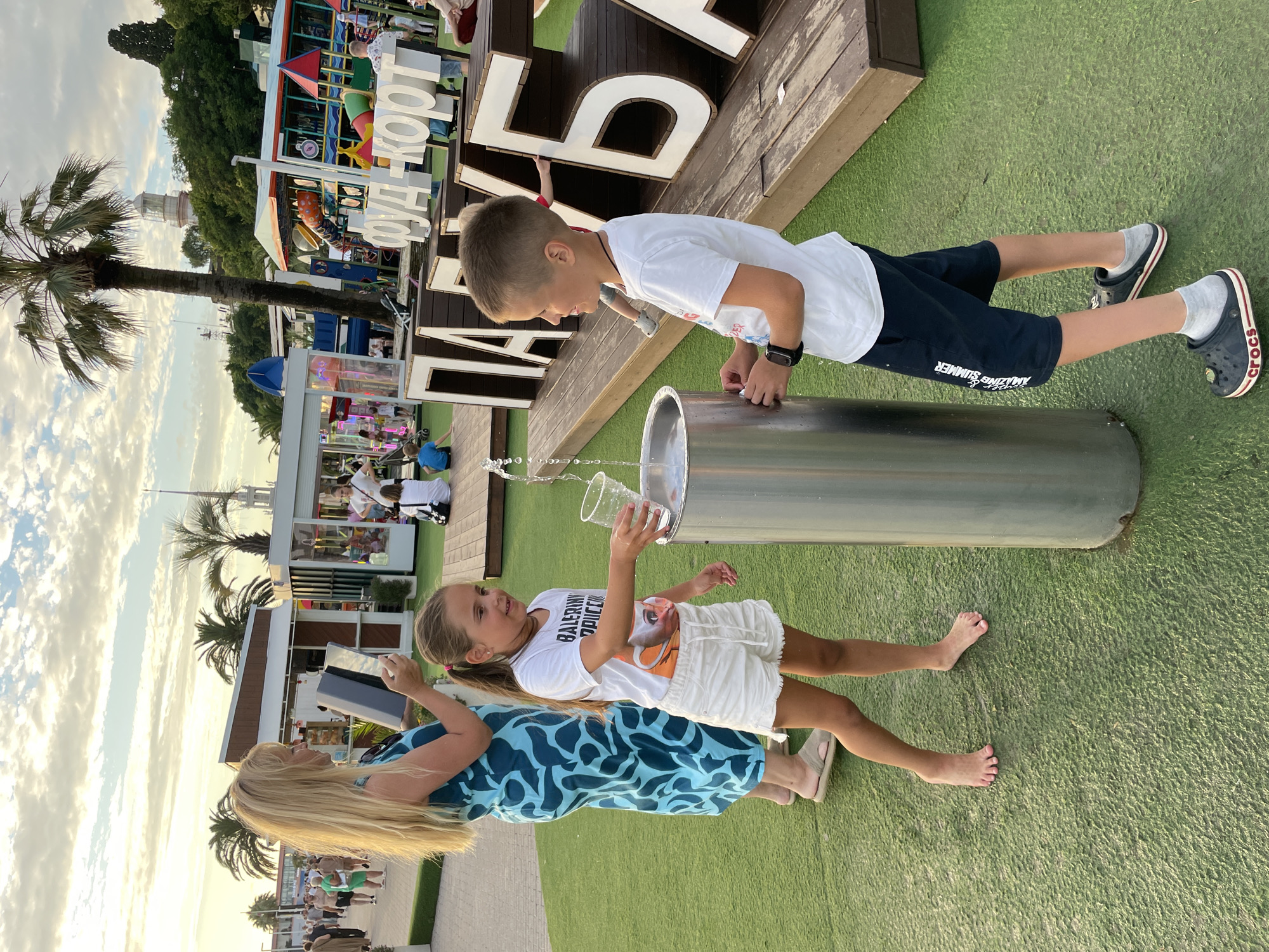 The image captures a sunny outdoor scene, likely in Sochi, Russia, given the presence of palm trees and a general resort-like atmosphere. In the foreground, a young girl with blonde hair pulled into a ponytail is holding a clear plastic cup under a fountain of water, seemingly filling it. She is wearing a white t-shirt and white shorts. Beside her, a young boy, also in a white t-shirt and dark shorts, is standing and looking towards the fountain. He appears to be holding something in his hand near the fountain. Behind them, a woman with long blonde hair, dressed in a blue patterned dress, holds a tablet device. The ground is covered in green artificial turf. In the background, there are various structures, including what appear to be food stalls or small shops, a playground with colorful slides, and large, decorative letters spelling out text in Russian, with "ПАРК" and "ФУД-КОРТ" visible. A lighthouse or tower is visible in the distance, atop a hill. People are seen walking in the background, suggesting a public recreational area. The sky is partly cloudy with soft lighting, indicating it might be late afternoon or early evening.