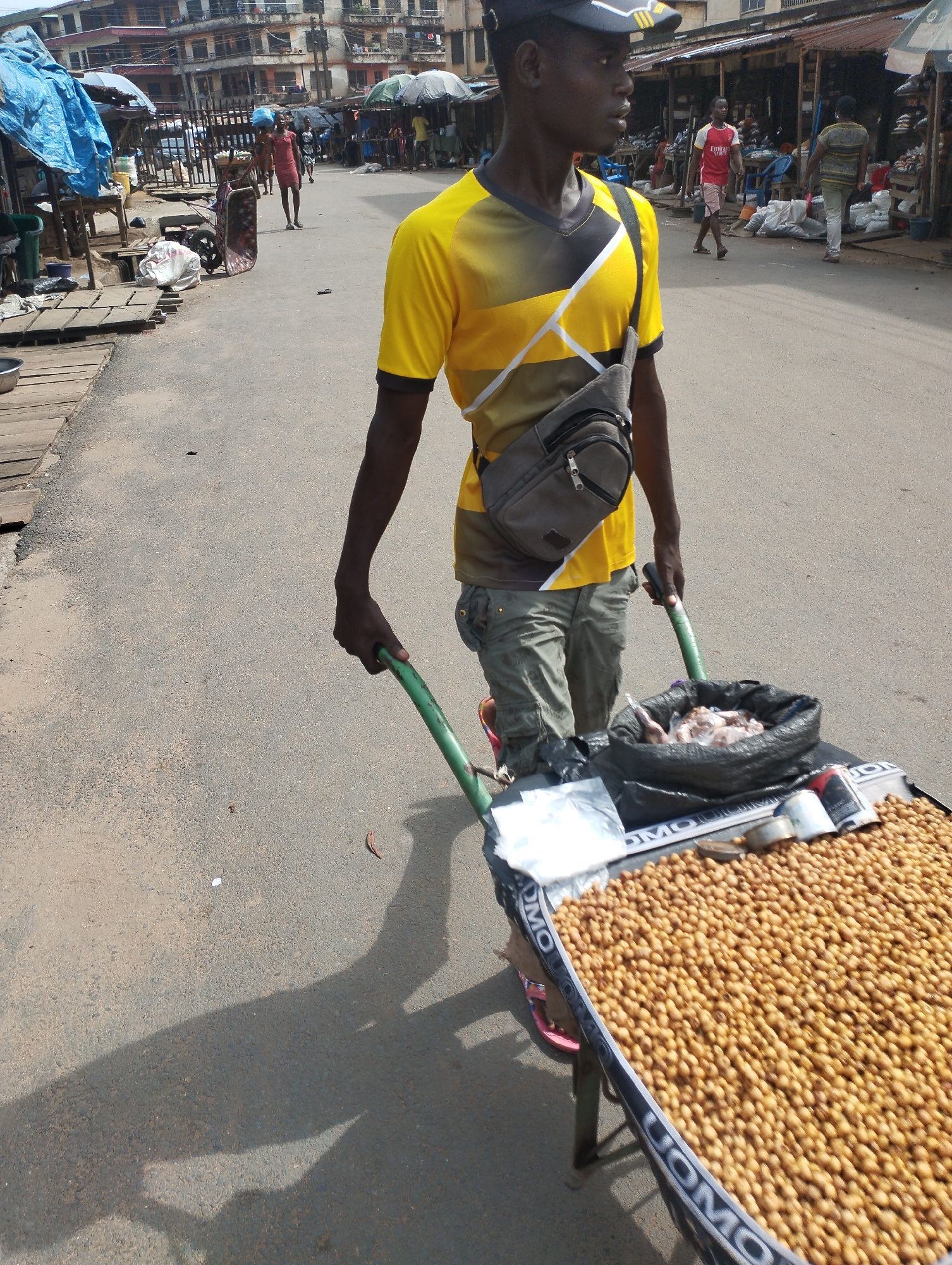 In Fegge, Nigeria, a man stands on a paved road pulling a green-handled, loaded wheelbarrow. He wears a yellow and black t-shirt, khaki pants, a dark cap, and a gray crossbody bag. The wheelbarrow is filled with a bed of small, round, tan food items and a black bag containing unidentified items. The edge of the wheelbarrow is labeled "UOMO". In the background, other people are walking along a street lined with buildings and market stalls shaded by umbrellas. The light suggests it is daytime.